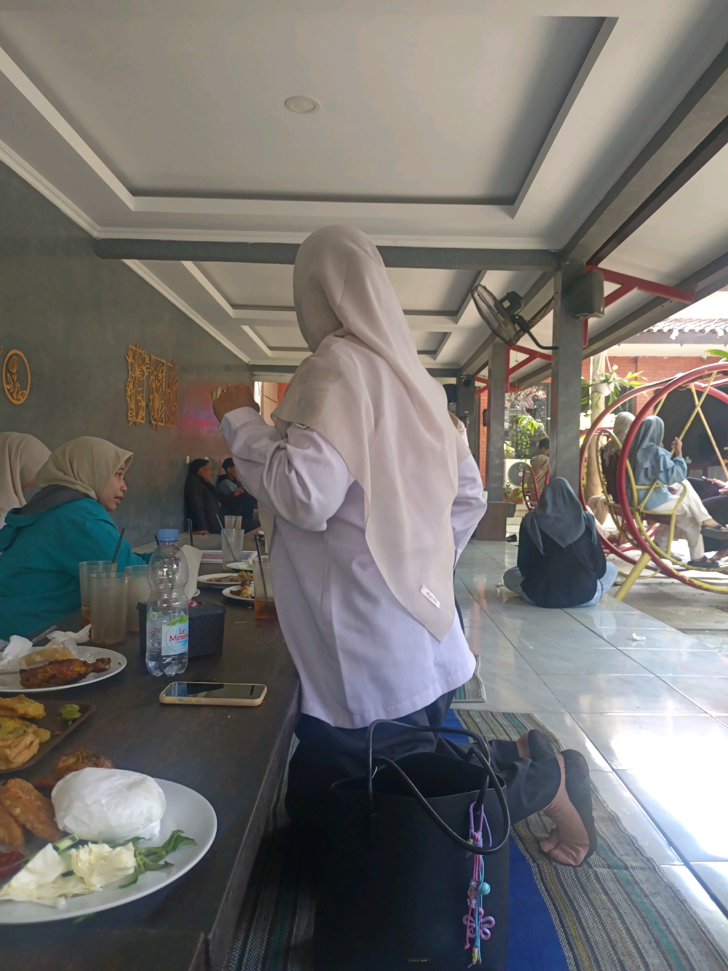 The image depicts an outdoor dining area in Kerangkulon, Indonesia, with several individuals visible. In the foreground, a person wearing a light-colored hijab and a white long-sleeved shirt is standing, facing away from the camera and towards a table laden with food and drinks. To the left, another person in a turquoise jacket and hijab is seated at the table, engaged in conversation. Other individuals are seated further back, some near a yellow and red circular swing set. The environment appears to be a covered patio with a tiled floor and a ceiling with recessed lighting. The table is set with plates of food, glasses, and a bottle of water. A black tote bag with colorful tassels rests on the floor next to the standing person. The overall scene suggests a casual gathering or meal.
