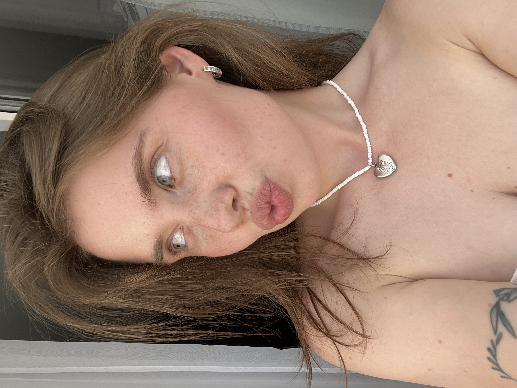 The image is a close-up, rotated selfie of a young woman. She has light brown hair that is slightly windswept. Her eyes are wide and looking to the side, with visible white in the irises. She is wearing a thin white beaded necklace with a heart-shaped pendant. The woman has a scattering of freckles across her nose and cheeks. Her lips are slightly parted and pursed. She is wearing a stud earring with small clear stones in her left ear. Her bare shoulder and upper chest are visible, and a tattoo of a leafy branch is visible on her upper right arm. The background appears to be indoors, with a sheer white curtain or fabric to the left and a dark surface behind her. The lighting suggests it is daytime. There is no visible text in the image. The location context of Chaikovsky, Russia is provided.