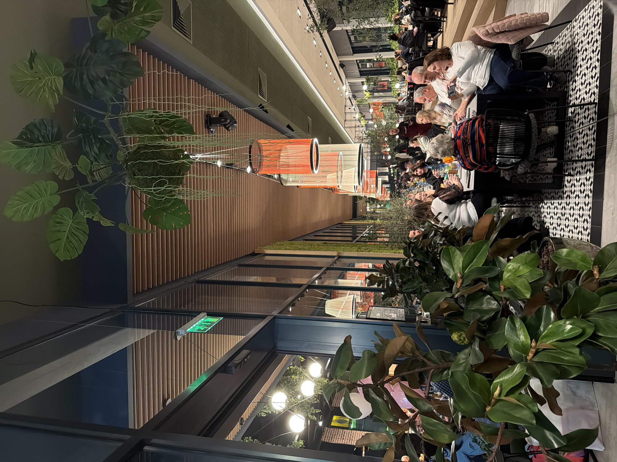 The image displays the interior of a contemporary dining establishment located in Plovdiv, Bulgaria. The perspective is directed down a long, well-lit hall occupied by numerous patrons. The ceiling features horizontal wooden slats interspersed with linear light fixtures and several large, decorative cylindrical pendant lights with orange and white woven textures. Artificial green foliage, including large-leafed plants and cascading vines, is suspended from the ceiling. A green illuminated "EXIT" sign is visible on a dark partition near the upper left.
The left wall comprises a tall glass partition reflecting the interior space. The floor is predominantly covered with black and white geometric patterned tiles, transitioning to a solid lighter color further into the venue. Large potted plants with dark green leaves are strategically positioned throughout, serving as decorative elements and subtle dividers.
In the mid-foreground, five individuals are seated around a dark square table. A person with grey hair is positioned with their back to the camera, wearing a multi-colored striped sweater. To their right, an older male with short grey hair is holding and looking at a white smartphone. Adjacent to him, an older female with brown hair is holding a white smartphone, pointing it towards the table, indicating active photography or video recording. Two other individuals, an older male and a female with dark hair, are partially visible at the table, engaged in interaction. The table holds several glasses containing beverages.
Numerous other patrons are visible seated at various tables further down the hall, suggesting a busy dining environment. A male staff member in a black t-shirt is observed standing in the middle distance, holding a tray. The overall illumination is provided by a combination of overhead recessed lights, linear fixtures, and decorative pendants.