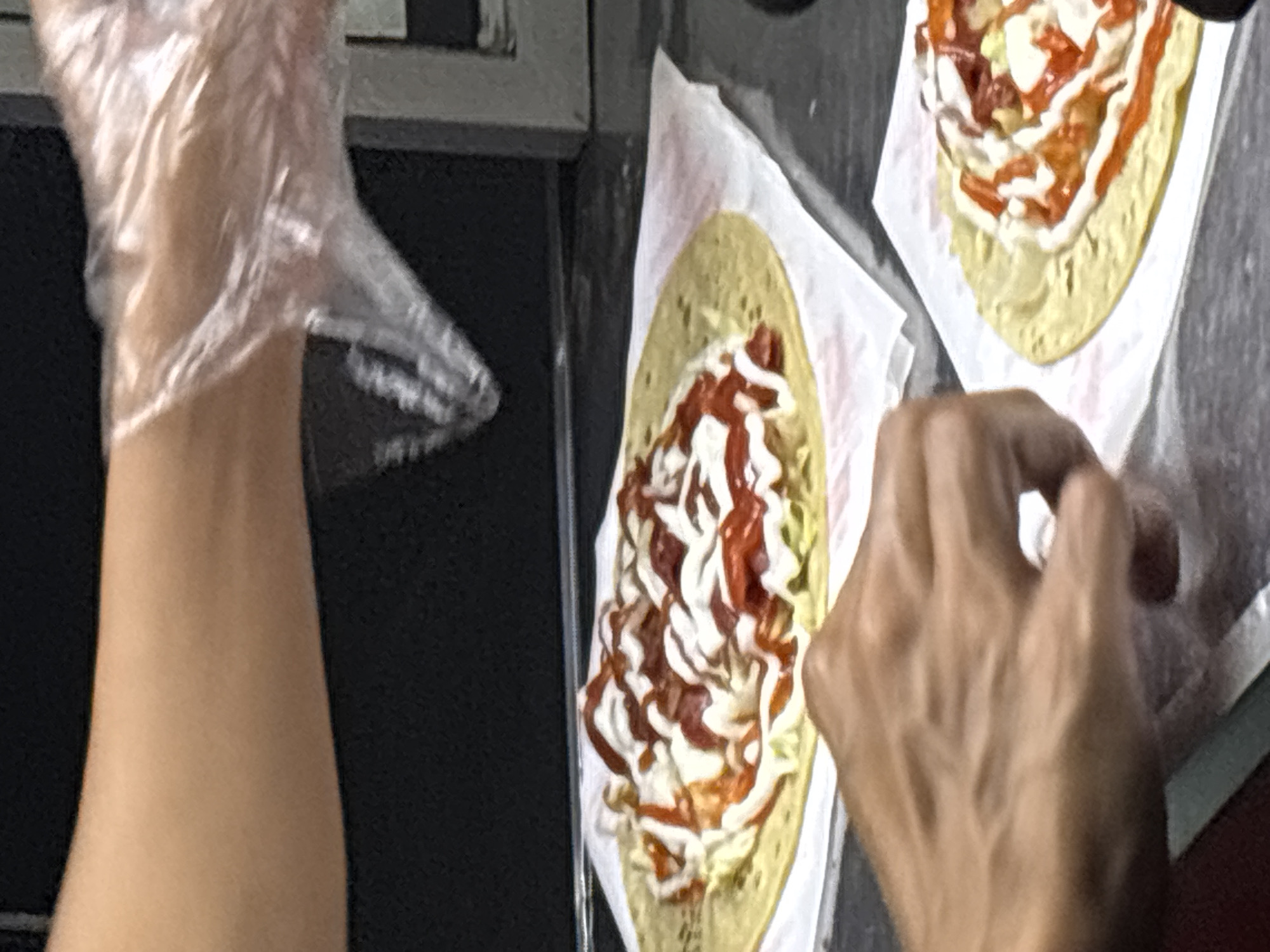 In North Jakarta, Indonesia, a close-up shot captures two prepared wraps with a hand reaching for one, suggesting a food service setting. The wraps, topped with a drizzle of white sauce and red sauce, are on white paper wrappers. One wrap is slightly out of focus in the foreground, with a hand in the lower left corner. The other wrap is in focus in the upper half of the frame, with a person's arm and hand wearing a clear plastic glove in the upper right, reaching over the wrap. The background is dark and indistinct, possibly indicating an indoor commercial environment.