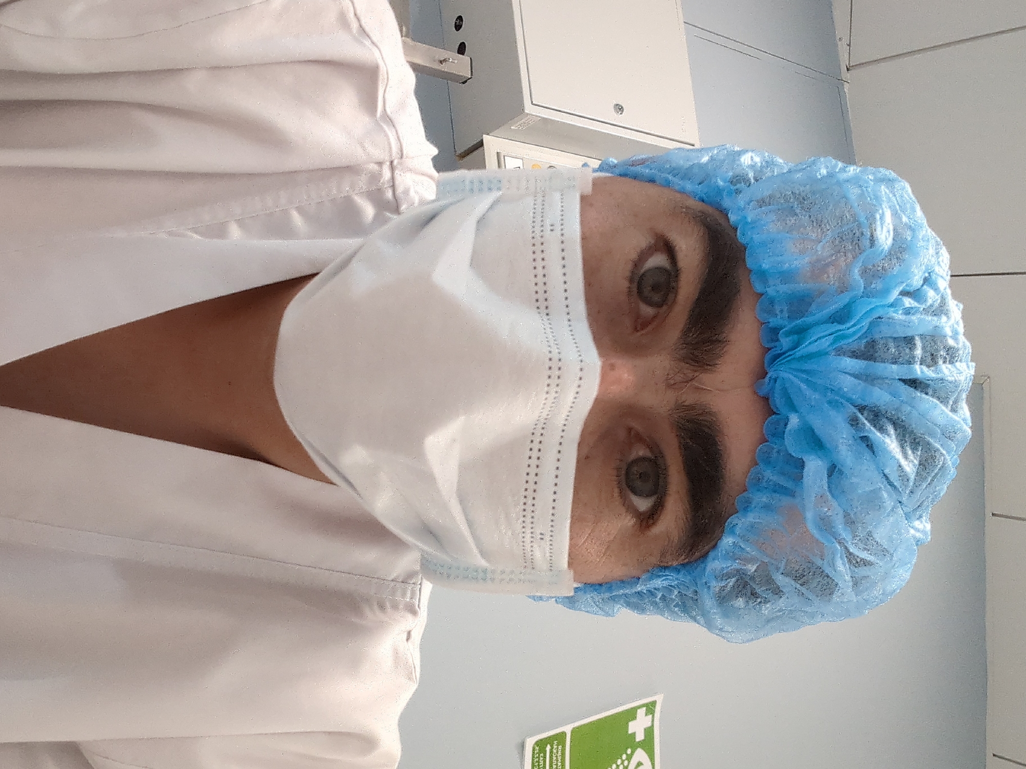 A close-up, eye-level selfie shows a person wearing a blue hair net and a white surgical mask, covering their nose and mouth. The person's eyes are visible above the mask. They are wearing a white lab coat. In the background, there is a white cabinet on the left, and a light blue wall with a green sign on the right. The sign has a white cross and some text in an unknown language. The overall lighting suggests an indoor environment, possibly a medical or laboratory setting. The image appears to have been taken in a city, but specific location details, such as Astana, Kazakhstan, are not visually discernible from the provided image alone.