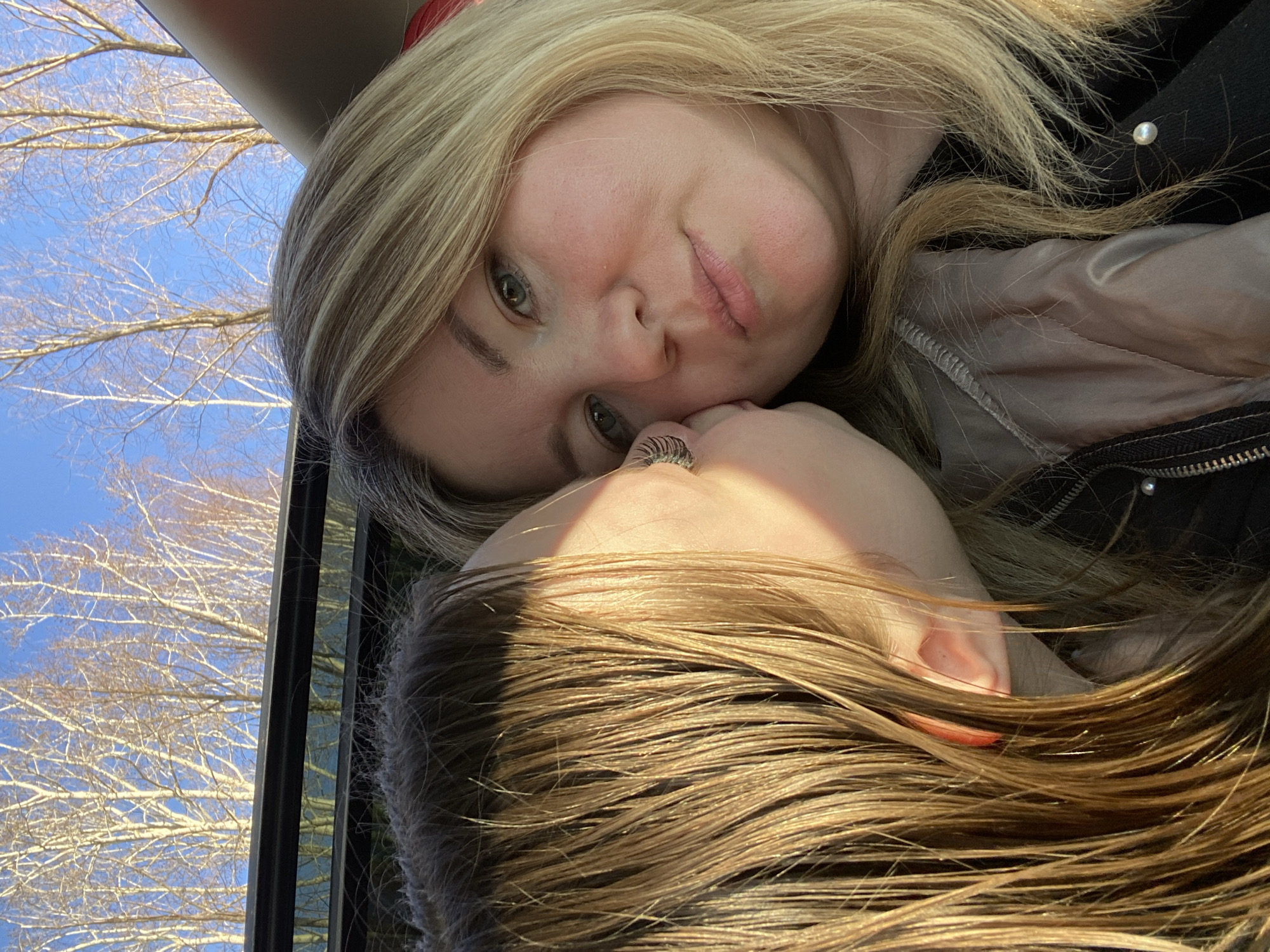 The image is a close-up, horizontally oriented shot featuring two individuals, likely women, in what appears to be a selfie-style composition. The person in the foreground has blonde hair and looks directly at the camera with a gentle, slightly puckered expression. Their fair skin shows a subtle blush, and they have light-colored eyes. A hand, wearing a silver-colored ring, is visible near the head of the second individual.
This second person has light brown hair and is positioned closely, with only the top of their head and a small part of their face or ear visible, mostly in shadow. Their hair catches highlights from a strong light source. The blonde person is wearing a dark top, possibly a jacket, with a small pearl-like embellishment on the shoulder.
Behind the subjects, a bright blue sky is visible through a window-like frame, along with the bare branches of deciduous trees. This suggests a clear day in late winter or early spring. The overall mood conveyed is intimate and warm. The scene is set in Chaikovsky, Russia. There is no visible text in the image.