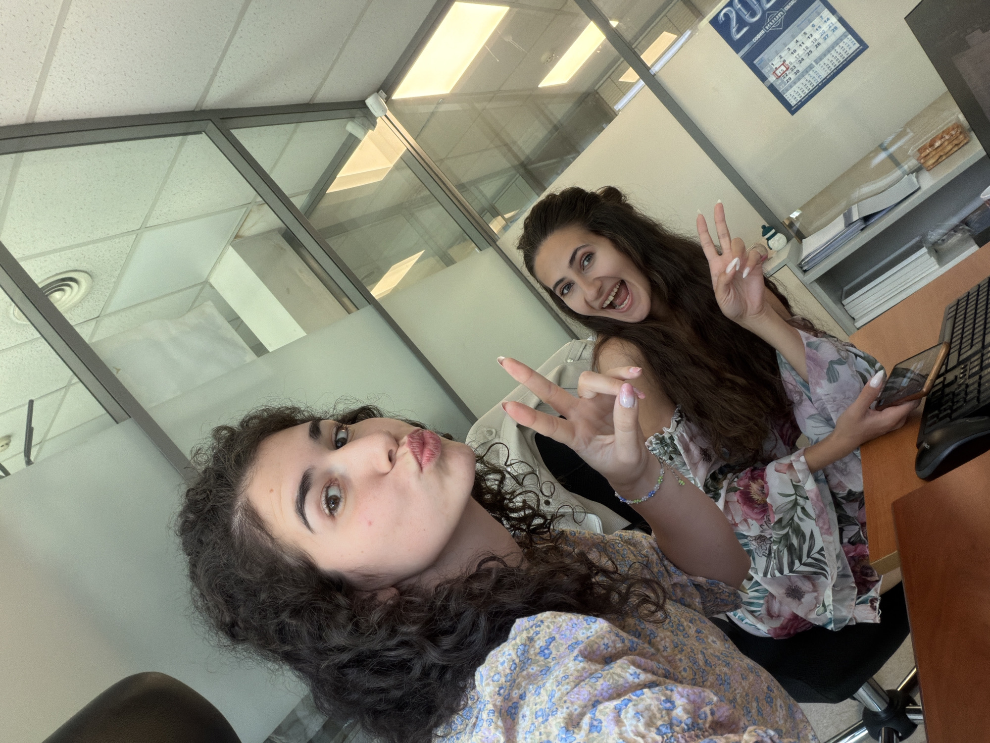 Two young women are taking a selfie in an office environment. The woman in the foreground on the left has dark, curly hair and is wearing a floral print top. She is puckering her lips and holding up two fingers in a peace sign. The woman on the right, with long brown hair, is wearing a floral print dress and is smiling broadly with her mouth open, also holding up two fingers in a peace sign. They are seated at desks, and behind them, large glass windows reveal a modern office ceiling with fluorescent lighting. A calendar for "2023" is visible on a wall in the background. The scene appears to be an office in Sofia, Bulgaria, judging by the style of the office and the calendar's display.