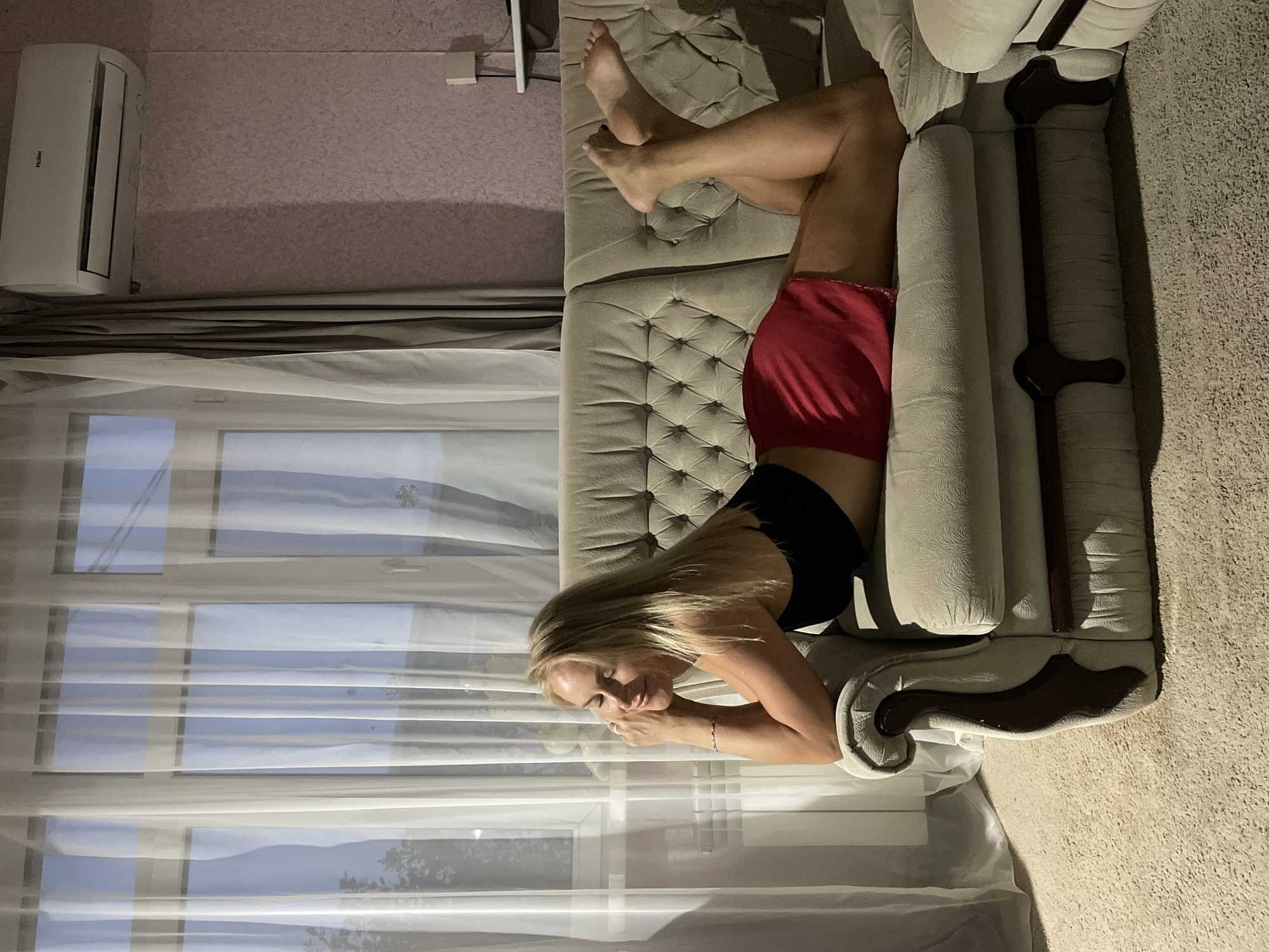 A woman is lying on a couch in a room. She is wearing a black top and red shorts. The couch is beige and upholstered. A window with sheer white curtains is behind her. An air conditioning unit is on the wall. The location is potentially a home or apartment in Baranovka, Russia, although this is not definitively confirmed in the image.