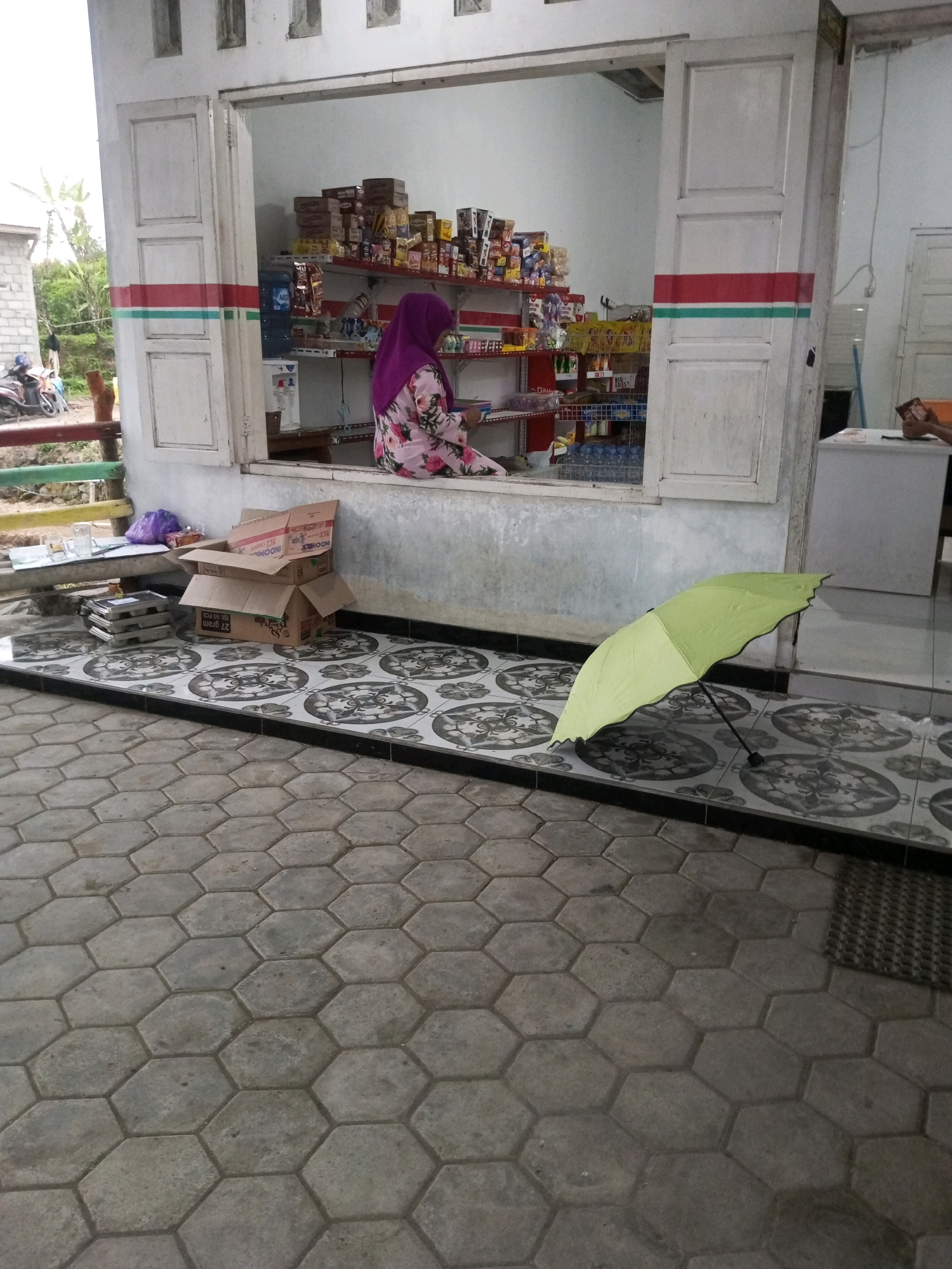 In Kalibeber, Indonesia, a scene unfolds outside a small shop. The shop, built with white walls and featuring two white doors with red and green horizontal stripes, has an open window where a woman wearing a purple hijab and a floral patterned dress is visible inside. She is seated and appears to be engaged with something on her lap, possibly a device. Shelves inside the shop are stocked with various products, including what appears to be food items and beverages. To the right of the shop, a white counter is visible.
Outside the shop, a light green umbrella is open and resting on a patterned tiled floor. Near the entrance, several cardboard boxes are stacked, with one box open and containing smaller boxes inside. To the left, there is a small table with glasses and a purple plastic bag. The ground outside the tiled area is paved with hexagonal concrete blocks. In the background, a motorbike is parked, and some greenery is visible, suggesting a semi-rural or suburban setting. The overall impression is of a small, local retail establishment in a residential or community area.