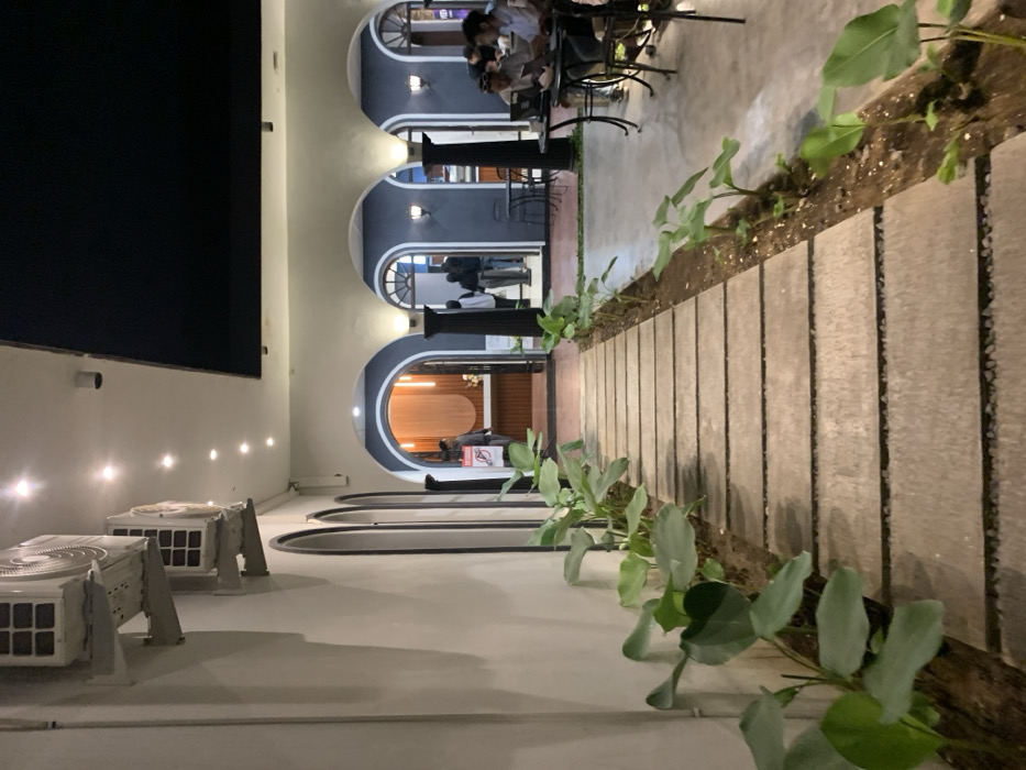 The image shows a walkway lined with large, green, leafy plants, leading to an entrance of a restaurant or cafe with arches. The entrance leads to a seating area with a few people seated at tables. The walkway is made of stone and leads to a courtyard. There are two large, white air conditioners on the wall of the building, which is painted white. The image was taken at a location in Malang, Indonesia with latitude -7.955 and longitude 112.634.
