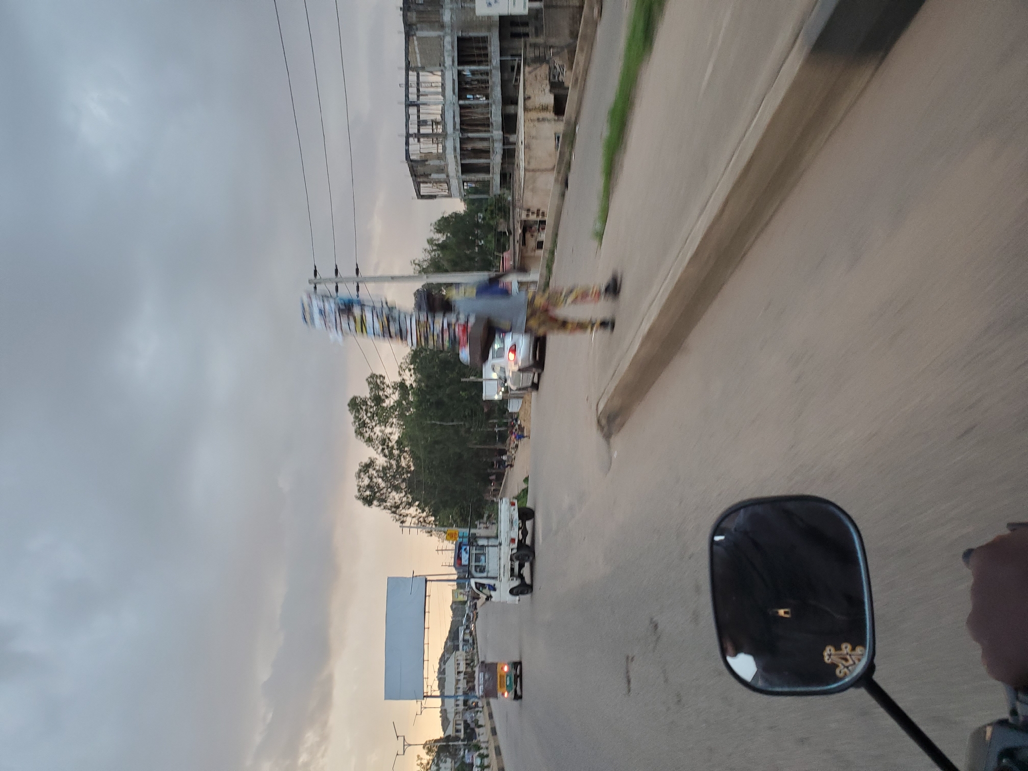 The image depicts a street scene in Gura Top, Nigeria, under an overcast sky. A person is walking along the sidewalk carrying a large, tall stack of what appear to be magazines or papers. The individual is dressed in patterned trousers and a grey shirt, and their movement is blurred, indicating they are in motion. Behind them, an unfinished building with exposed concrete and rebar stands prominently.
In the foreground, a motorcycle's rearview mirror is visible, reflecting the rider's dark jacket and a metallic pendant. The motorcycle is in motion, indicated by the blur of the road.
On the road, a white pickup truck is driving away from the camera. Further ahead, a yellow motorized tricycle is also moving away. Other vehicles and a large, blank billboard structure are visible in the background, along with trees and buildings lining the street. The overall atmosphere suggests a moderately busy urban environment.