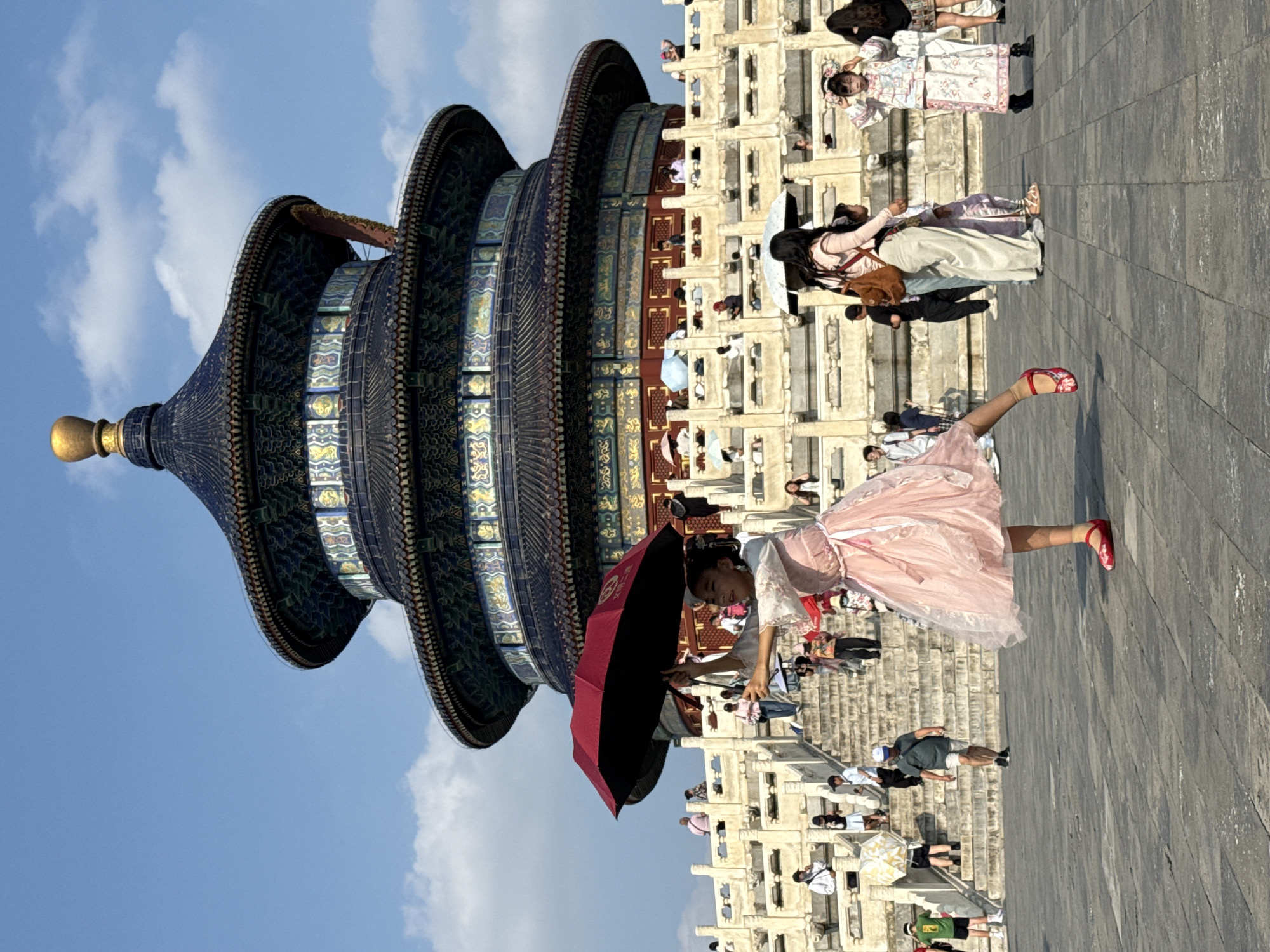 An outdoor, eye-level, medium-wide shot captures the Hall of Prayer for Good Harvests in the Temple of Heaven, Dongcheng District, China, under a clear blue sky with scattered white clouds. A young girl in a pink, layered dress and red shoes is the prominent subject in the foreground, holding a partially opened red umbrella above her head. Her left leg is extended upwards and forward, her right leg bent, and her body is turned to her right, suggesting a dance-like pose.
Behind her, a large multi-tiered, ornate temple structure dominates the background, its dark blue, multi-layered roof adorned with gold finials. White stone balustrades and a long flight of stairs lead up to the temple, with numerous people scattered throughout the scene. Some individuals are ascending the stairs, while others are standing on the platforms, many holding umbrellas to shield themselves from the sun. The ground in the foreground is paved with large, grey stone tiles. The lighting suggests it is a sunny day.