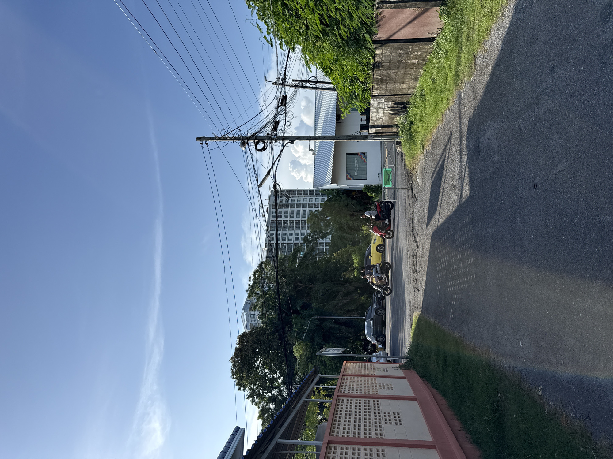 In an urban environment, on a paved road, a motorcycle and a scooter are visible. The motorcycle is red and is being ridden by a person in a white shirt. The scooter is black and gray, with two individuals on it, one driving and the other a passenger. Behind them, a yellow car and a dark-colored sedan are also present on the road. The surrounding area features a building with a white facade and a red-trimmed wall with a patterned design. To the right, a concrete wall borders lush green foliage and a tall building with multiple floors and balconies is visible in the distance. Overhead, a complex network of electrical wires is suspended from utility poles against a clear blue sky with wispy clouds. The scene suggests a typical street in Phuket City Municipality, Thailand, likely during daylight hours given the bright lighting and shadows.