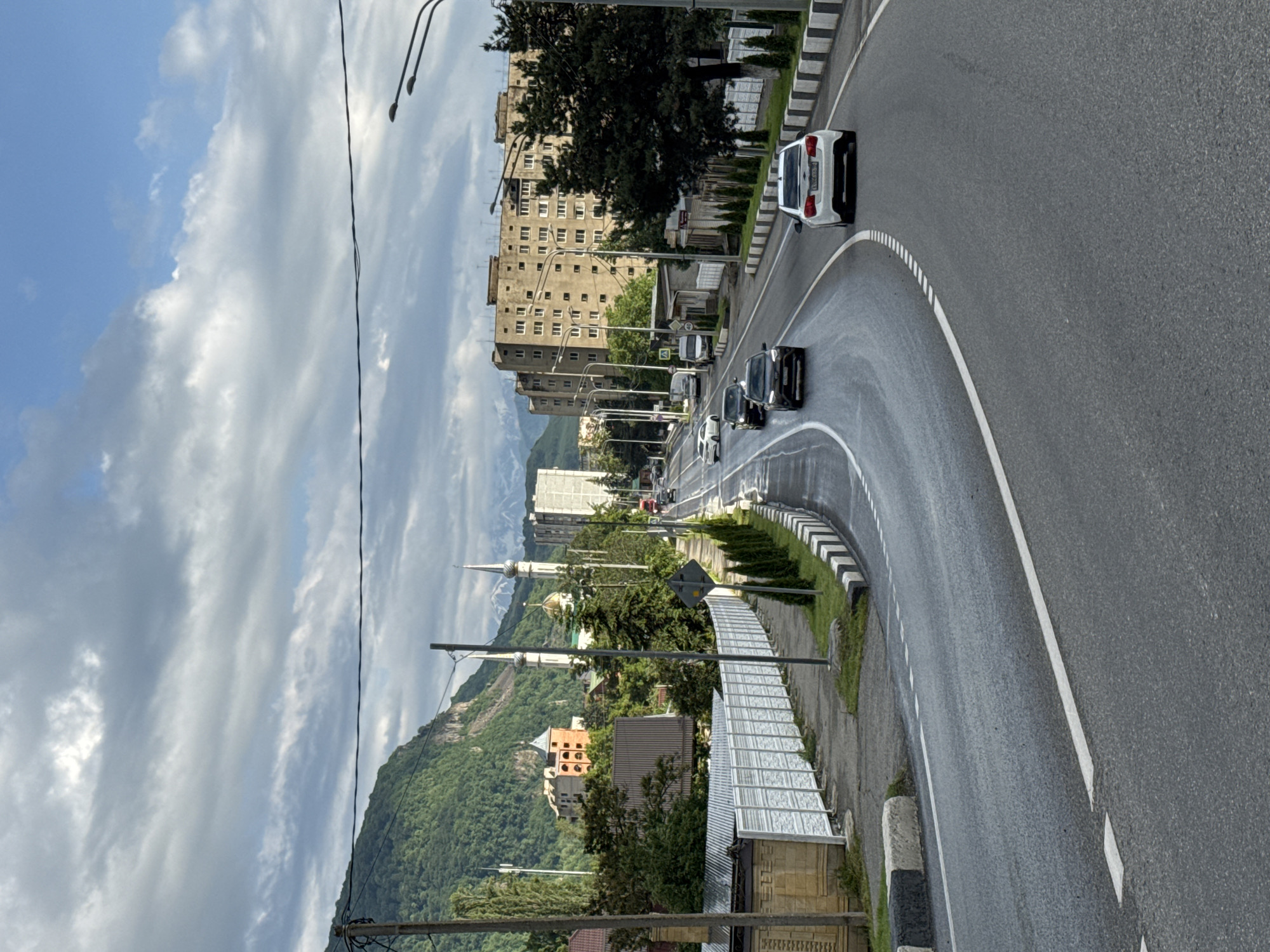 The image captures a wide, wet asphalt road in Karachayevsk, Russia, curving through an urban landscape with a mountainous backdrop. The road is marked with white lane dividers and bordered by a black and white striped curb. Several vehicles are visible on the road, including a white sedan in the foreground, a dark SUV, and other cars further down. On the left side of the road, there is a paved sidewalk, a low white fence, and various buildings, including what appears to be a mosque with minarets and a dome, and an orange brick building with a distinctive turreted roof. Further in the background, a large, multi-story concrete apartment building dominates the skyline, with other structures and trees interspersed. Lush green mountains, partially obscured by clouds, rise dramatically behind the city. The sky is a mix of blue and white clouds. The scene appears to be a typical urban street with natural mountainous scenery.