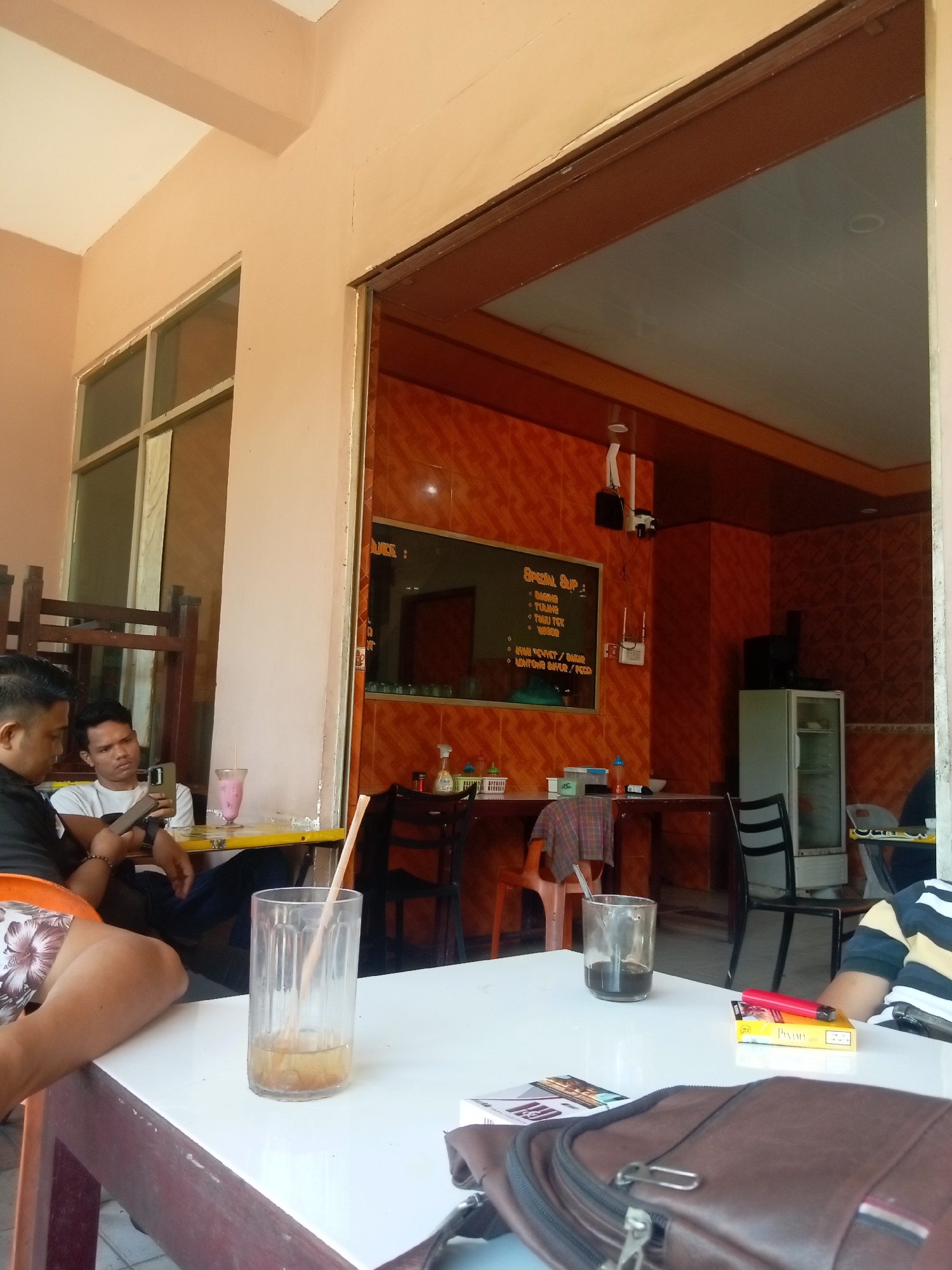 The image depicts an outdoor seating area of a cafe in Batam, Indonesia. In the foreground, a white table is set with several items: a glass with a straw and some liquid, a pack of cigarettes, a red electronic cigarette device, a wallet, and a brown bag. To the left, two men are seated at a yellow table. One man, wearing a black shirt and patterned shorts, is looking at his phone. The other man, in a white t-shirt, is also looking at his phone. A pink beverage is on the table in front of him. In the background, the interior of the cafe is visible, featuring orange tiled walls, tables with chairs, a refrigerator, and a menu board. The overall scene suggests a casual setting where people are relaxing and socializing.