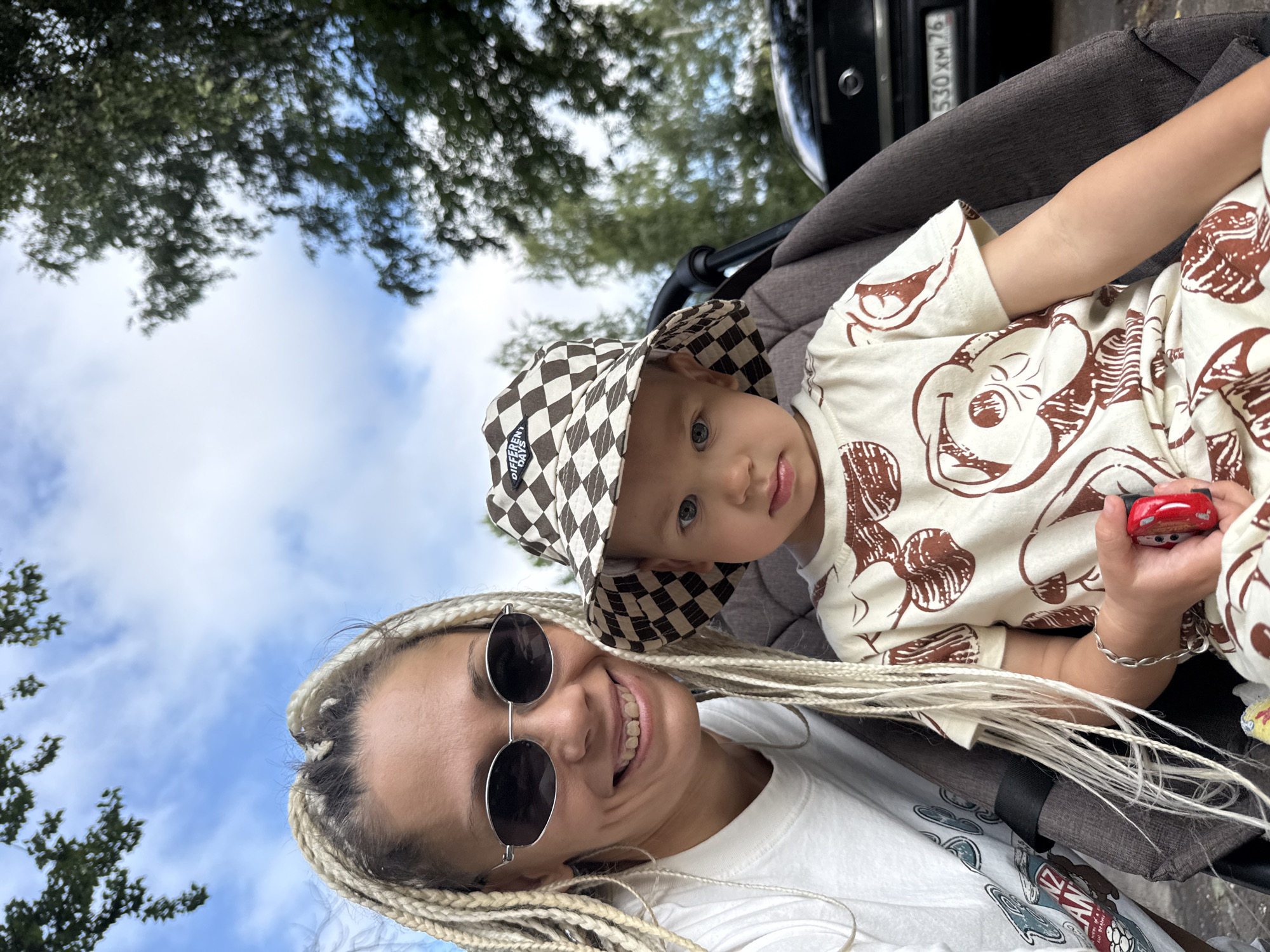 In Rusanovo, Russia, a woman with blonde braids and sunglasses smiles at the camera, taking a selfie with a young child in a stroller. The child, wearing a checkered hat and a patterned shirt, looks directly at the viewer. The background features trees and a car with a license plate reading "530 XM 76." The sky is blue with scattered clouds.