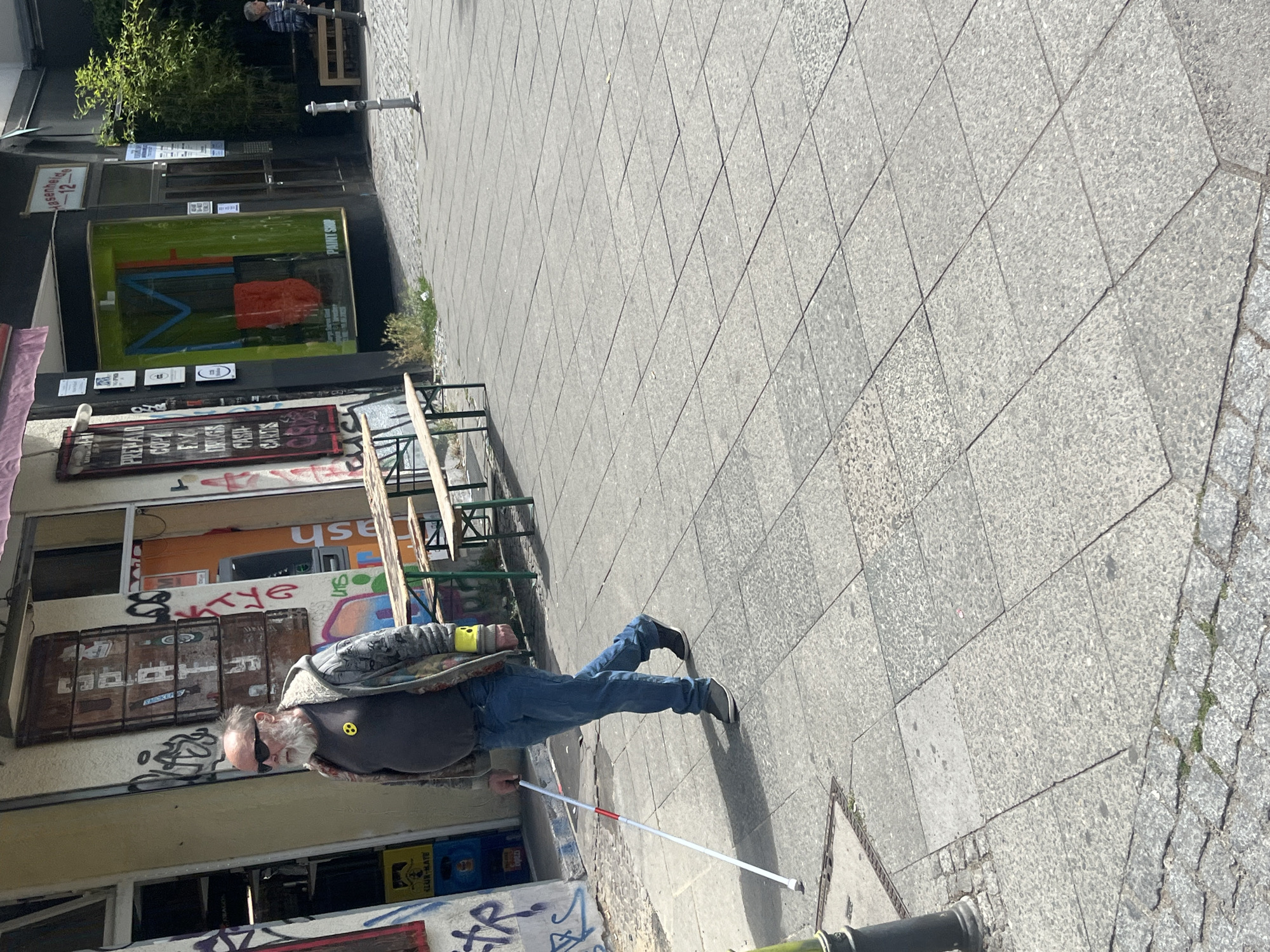 In Berlin, Germany, a man with a white beard and sunglasses walks on a sidewalk holding a white cane. He is wearing a grey and patterned jacket over a dark t-shirt with a yellow smiley face logo. He has on blue jeans and dark shoes. The sidewalk is paved with large grey tiles. Behind the man, there is a building with graffiti on its facade. Several wooden picnic tables are set up in front of the building. Further down the street, there is a shop with a green facade and a brightly colored display visible through the window. A few trees are planted along the street. The scene appears to be a public street with shops and outdoor seating.
