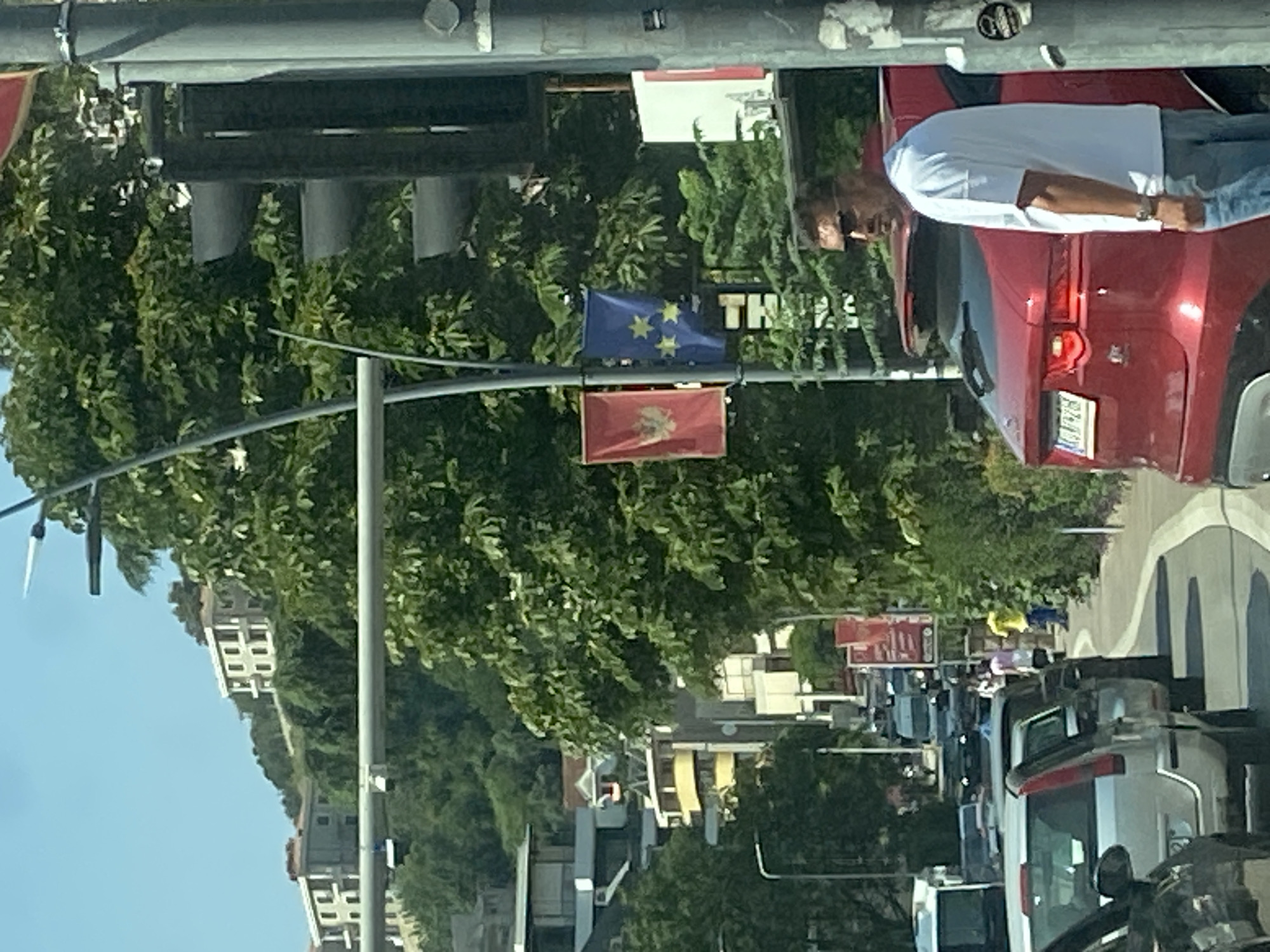 The image captures a traffic scene in Budva, Montenegro, on a sunny day. In the foreground, a red Jaguar F-Pace SUV is parked, with a man wearing a white t-shirt and blue jeans standing beside it, looking to his right. The SUV has a license plate that appears to be from Montenegro. Behind the SUV, several cars are lined up in traffic, suggesting a congested road.
Further back, a pedestrian walkway is visible, with a few people walking. The environment is characterized by lush green trees and buildings on a hillside, typical of Budva's urban landscape. Two flags are prominently displayed on a lamppost: one red with a golden eagle emblem, likely the flag of Montenegro, and another blue flag with yellow stars, representing the European Union. The presence of these flags might indicate an event or a specific location of significance. The background shows residential buildings scattered on the hilly terrain, bathed in bright sunlight.