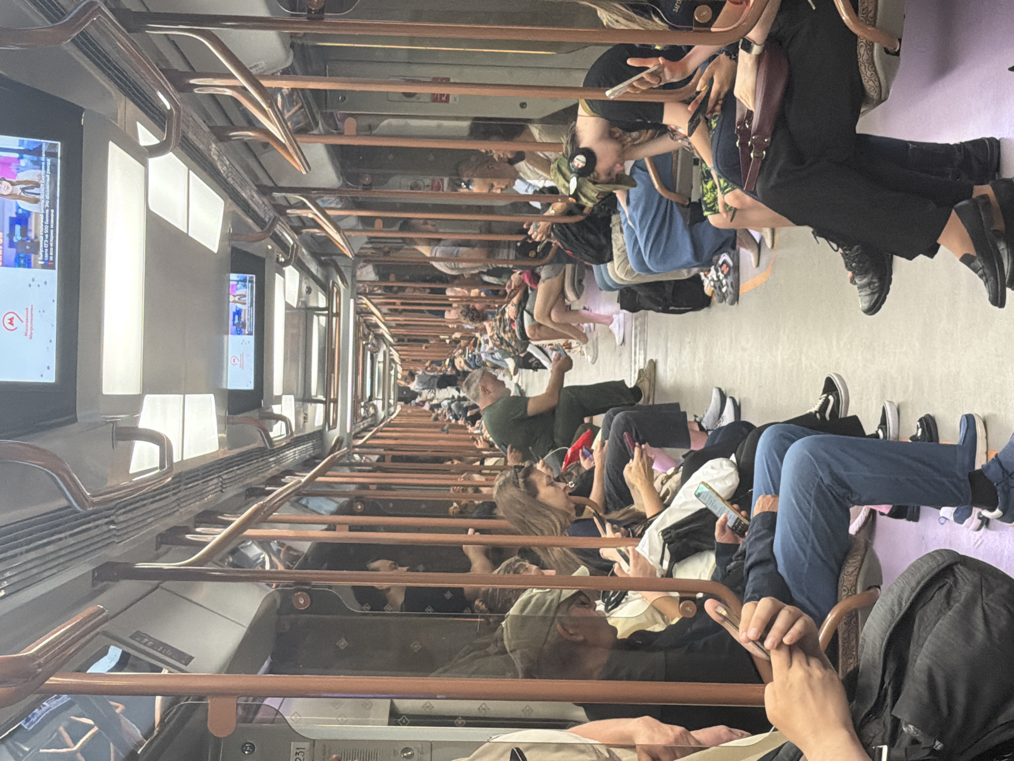 The image shows the interior of a subway car with many passengers seated. The car is illuminated by overhead lights and has orange handrails running along the ceiling and sides. The seats are upholstered in a patterned fabric. Most passengers are looking at their phones. In the foreground, a person wearing a dark jacket and blue pants is holding a phone. To their right, a person in black pants and shoes is holding a phone. In the middle ground, a man in a green shirt is seated, also looking at his phone. Further down the car, more passengers are visible, some appearing to be sleeping or resting. There are advertisements displayed on screens at the front of the car. The overall impression is one of a typical commute in an urban subway system.