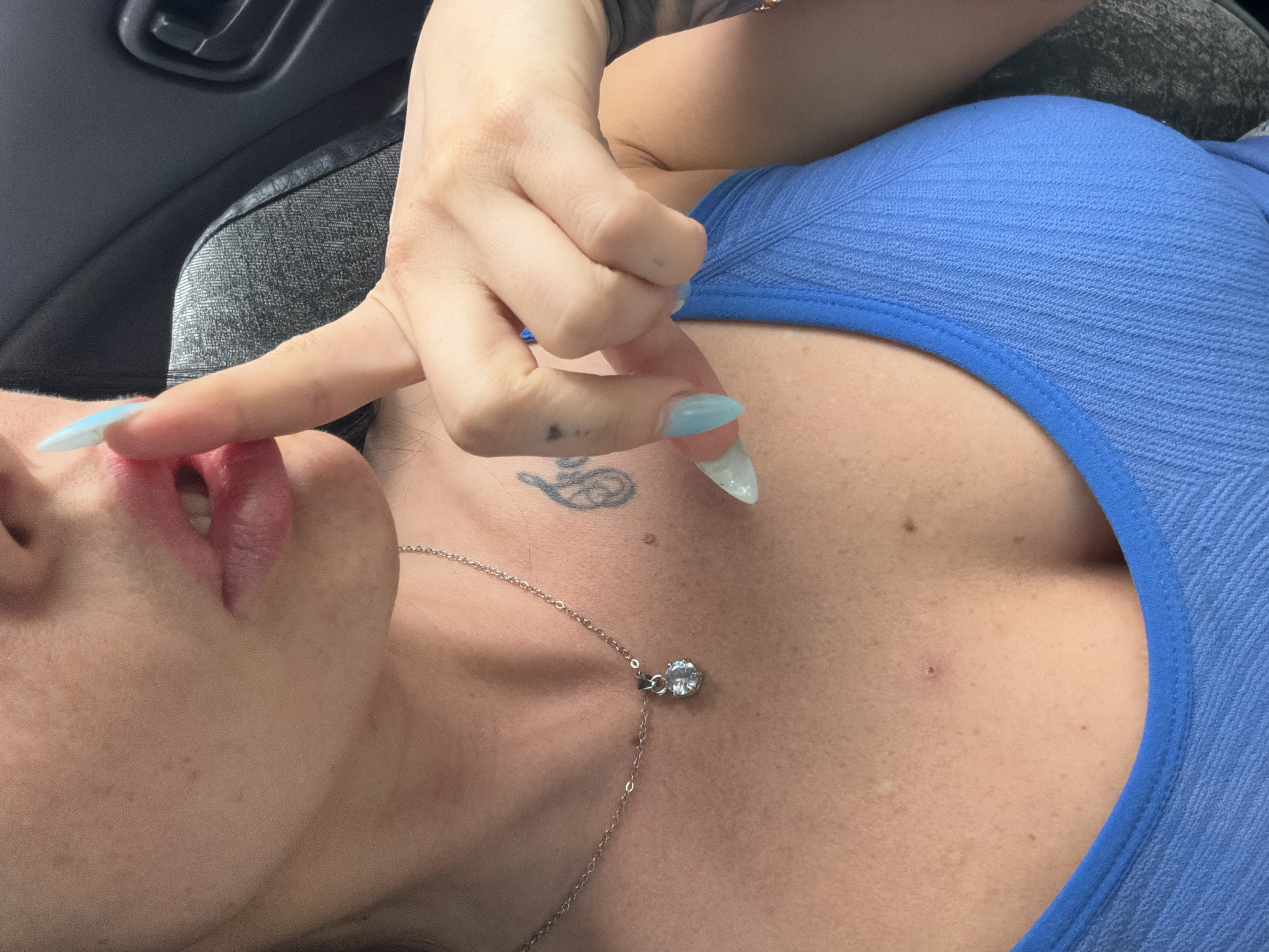 The image shows a close-up view of a person's chest and face. The person is wearing a blue, textured top. A silver necklace with a clear pendant is visible on their chest. The person has a tattoo on their upper chest, and their fingernails are long and appear to be painted a light blue color. One finger is touching their lips. The background suggests an indoor setting, possibly a car, with a dark gray handle and seating visible. There is no text visible in the image. The lighting suggests it is daytime.