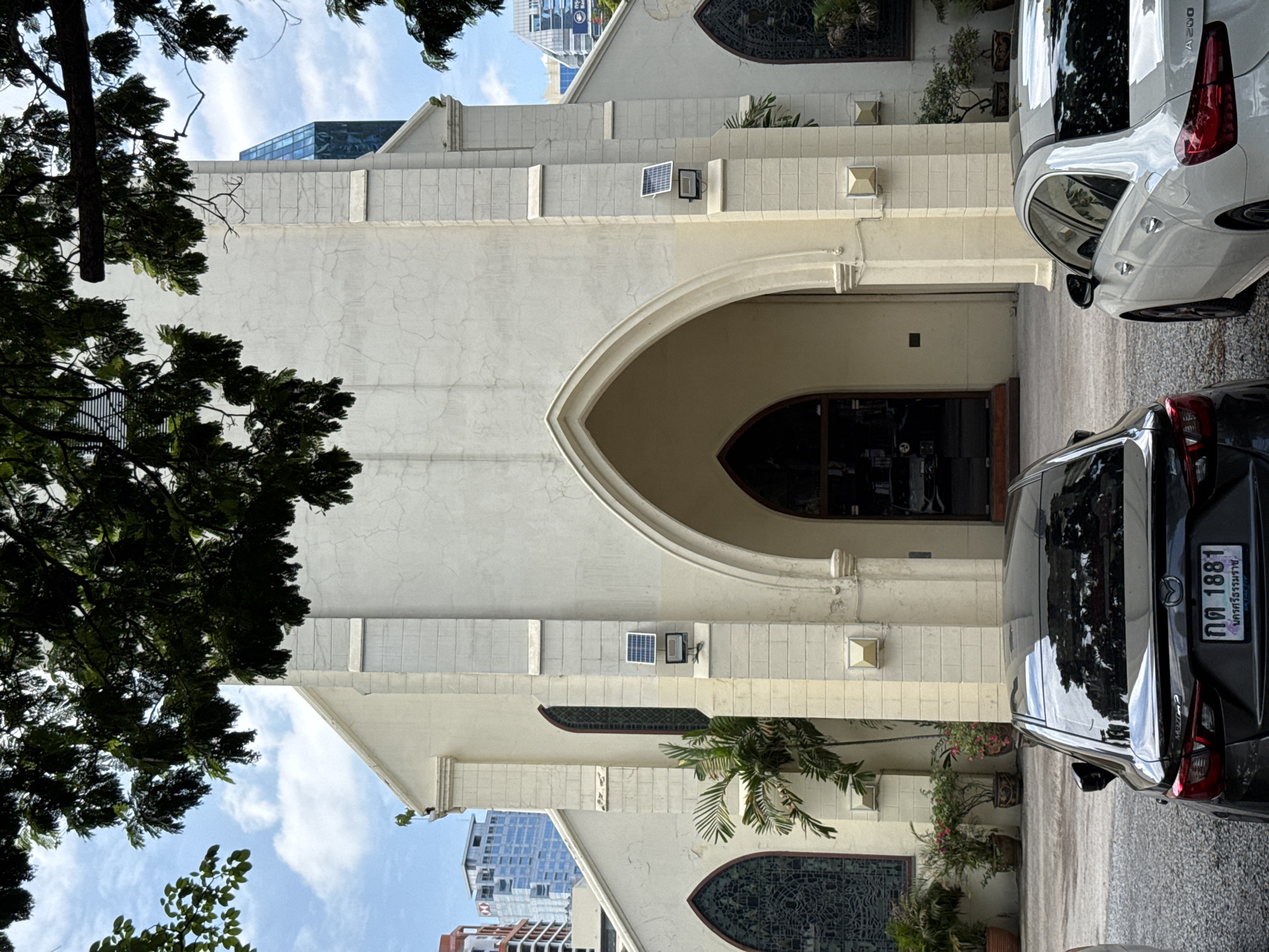 The image shows the facade of a light-colored church in Bangkok, Thailand, framed by dark green foliage from trees in the foreground. The church has a prominent arched entrance, flanked by two tall, narrow arched windows with stained glass. Above these windows, on the exterior wall, are two solar panels with attached lights. The building's architecture features decorative stone elements and pilasters. In the background, modern city buildings, including skyscrapers, are visible, indicating an urban setting. In front of the church, two cars are parked on a gravel surface. The car closer to the viewer is black and the other is white. The sky is partly cloudy.