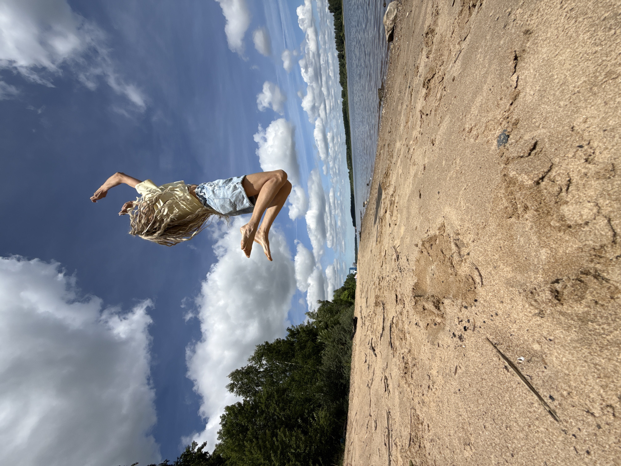 The image captures a person suspended mid-air against a bright blue sky, suggesting a jump, flip, or playful moment. The individual has long, curly blonde hair that is fanned out due to the motion, and is wearing a light yellow short-sleeved top and blue denim shorts. Their bare arms are outstretched, and bare feet are visible.
The scene is set outdoors on a sunny day, likely on a sandy beach. The foreground features textured brown sand, which appears to be a shoreline. To the right, a calm body of water, possibly a lake or a wide river, stretches towards the horizon. A distant line of lush green trees marks the far bank of the water. The sky is predominantly blue with scattered, fluffy white cumulus clouds. The low-angle perspective emphasizes the person's height in the air and the vastness of the sky.
The activity depicted is a dynamic leap or acrobatic pose, conveying a sense of energy and freedom. The weather is clear and bright. The location, given the context, is Rusanovo, Russia, indicative of a natural, lakeside or riverside environment. There is no visible text in the image.