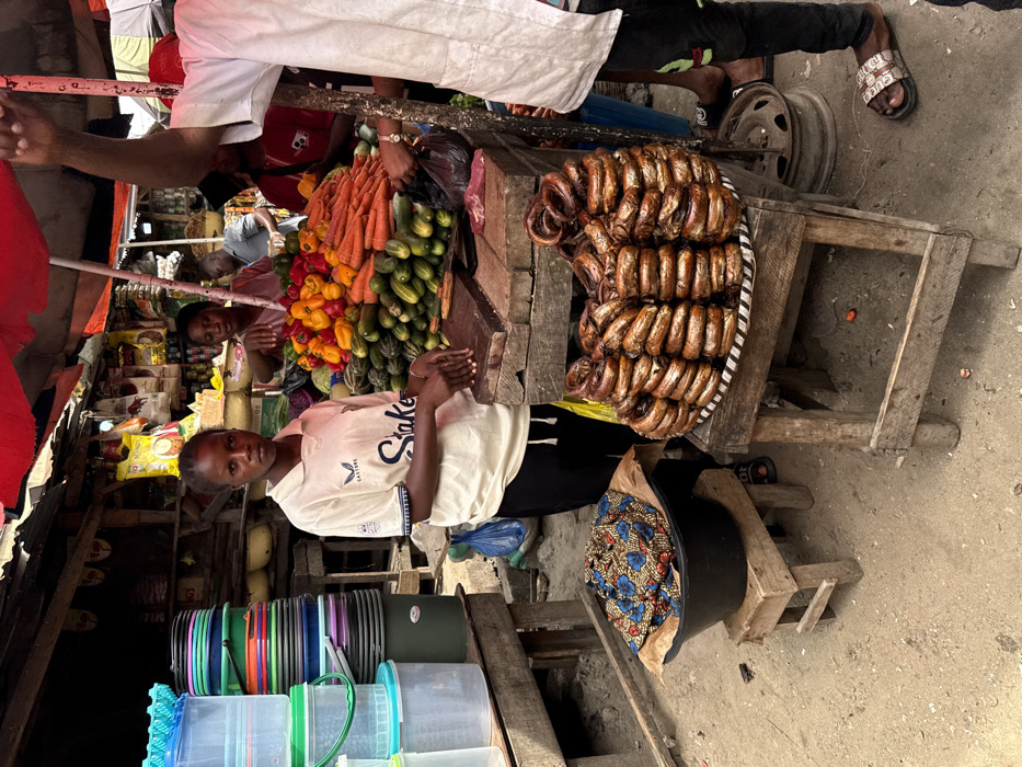 The image captures a vibrant, bustling scene within an outdoor market, likely during daytime hours. The perspective is slightly elevated, looking down into the market stalls, which are constructed from simple wooden frames.
In the center of the foreground, a young person, possibly male, stands facing the camera with a calm, neutral expression. He is wearing a light-colored, short-sleeved t-shirt with "Skake" (or "Snake") and "Castore" visible on the front. His hands are clasped together at his waist. To his left, another individual is partially visible, also looking towards the camera with hands clasped.
Behind these individuals, two main market stalls are prominently displayed. The stall on the left is piled high with a colorful array of fresh produce, including bright orange carrots, green cucumbers or zucchini, and a heap of red, yellow, and orange bell peppers. Adjacent to this vegetable stall, on the right, a large, round tray or woven mat is laden with numerous pieces of what appear to be smoked or dried fish, possibly eels, uniformly coiled into rings, presenting a dark, glossy, reddish-brown appearance.
Further back and slightly above the vegetable stall, a person dressed in a white top or robe and black trousers is partially visible, with their hands reaching into a black bag, suggesting they are handling merchandise or making a transaction. Their feet are visible, wearing sandals that appear to have some text on them. A metal wheel rim rests on the ground beneath their leg.
To the far left of the frame, a variety of household items are stacked. These include colorful plastic buckets in shades of green, blue, orange, and black, alongside clear plastic containers with colored lids. On the ground, near the dried fish stall, a large black basin or pot holds folded fabric patterned with blue, red, and white floral designs. The ground surface throughout the market area is a mix of dirt and worn concrete. In the background, behind the primary stalls, glimpses of more market activity and assorted goods can be seen, though less distinctly.