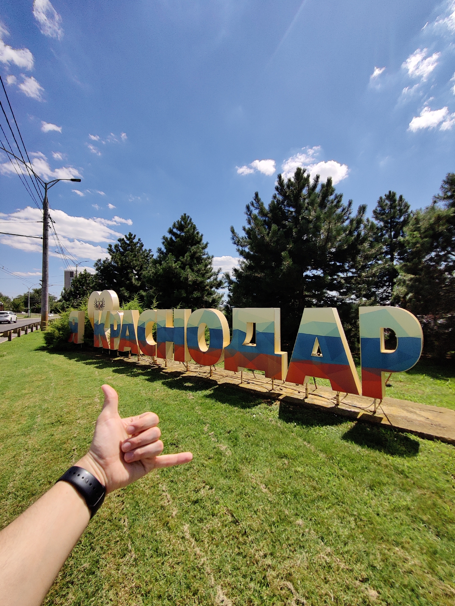 A low-angle, full shot captures a large sign spelling out "Я КРАСНОДАР" (Ya Krasnodar) in bold, three-dimensional letters with a Russian flag color scheme, set against a backdrop of green grass, trees, and a bright blue sky with scattered clouds. The sign appears to be made of wood or a similar material, with each letter supported by metal poles. In the foreground, a person's left arm and hand are visible, with the hand making a "shaka" gesture. The person is wearing a black smartwatch on their left wrist. To the left of the sign, a utility pole with wires and a streetlamp stands near a road where a white car is driving. The environment suggests a warm, sunny day in an outdoor public space in Krasnodar, Russia.