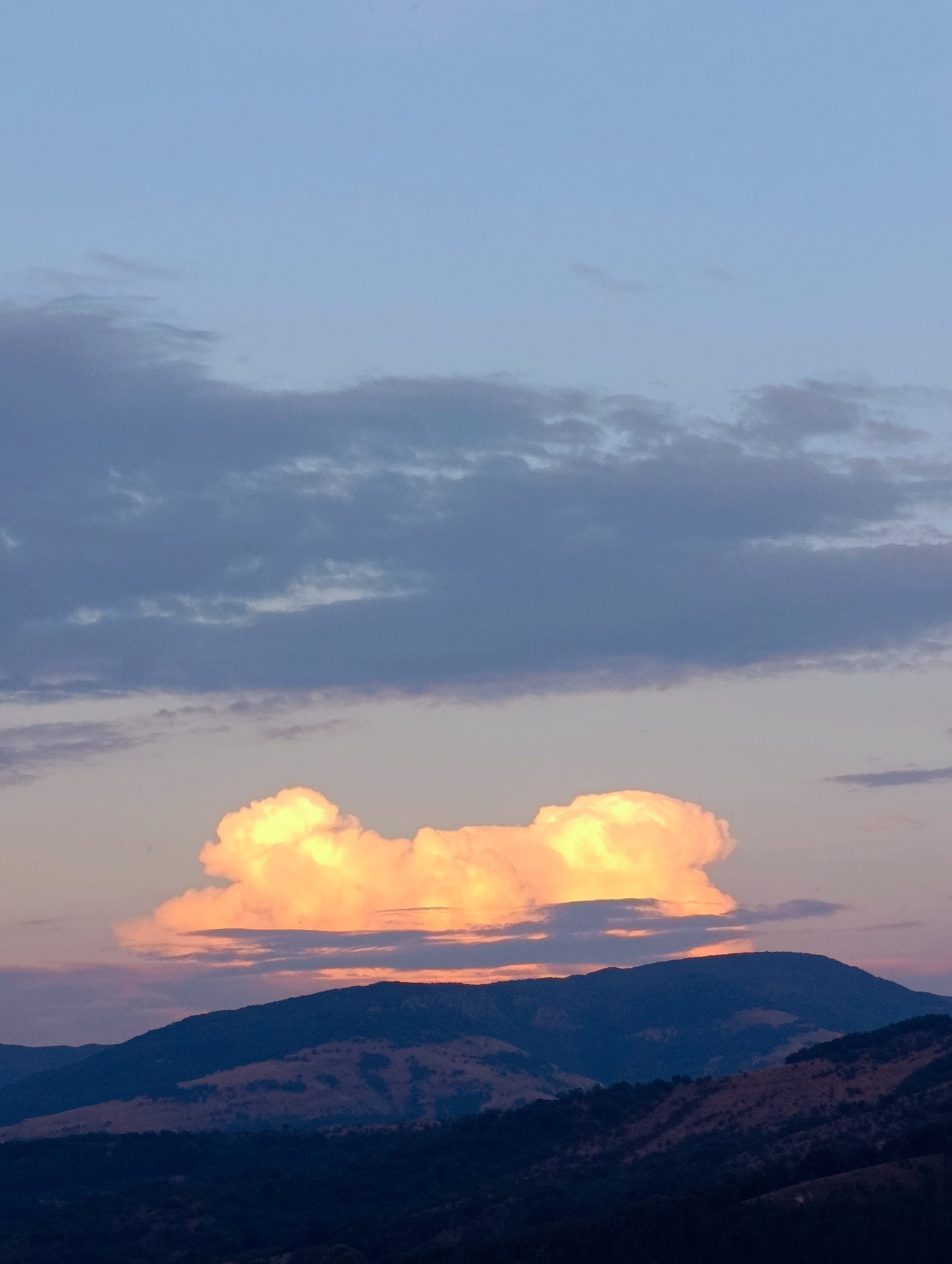 The image depicts a dramatic sunset over the hills near Shiroko Pole, Bulgaria. The sky is a gradient of soft blues and purples, transitioning to hues of orange and pink near the horizon. A large, cumulus cloud, illuminated by the setting sun, dominates the central portion of the sky. This cloud is a vibrant orange and yellow, casting a warm glow. Below the cloud, the silhouettes of rolling hills are visible, their dark forms contrasting with the colorful sky. The landscape appears rugged, with some areas showing patches of dried vegetation. The overall scene is one of natural beauty and tranquility, capturing a moment of intense color in the sky as day turns to night.