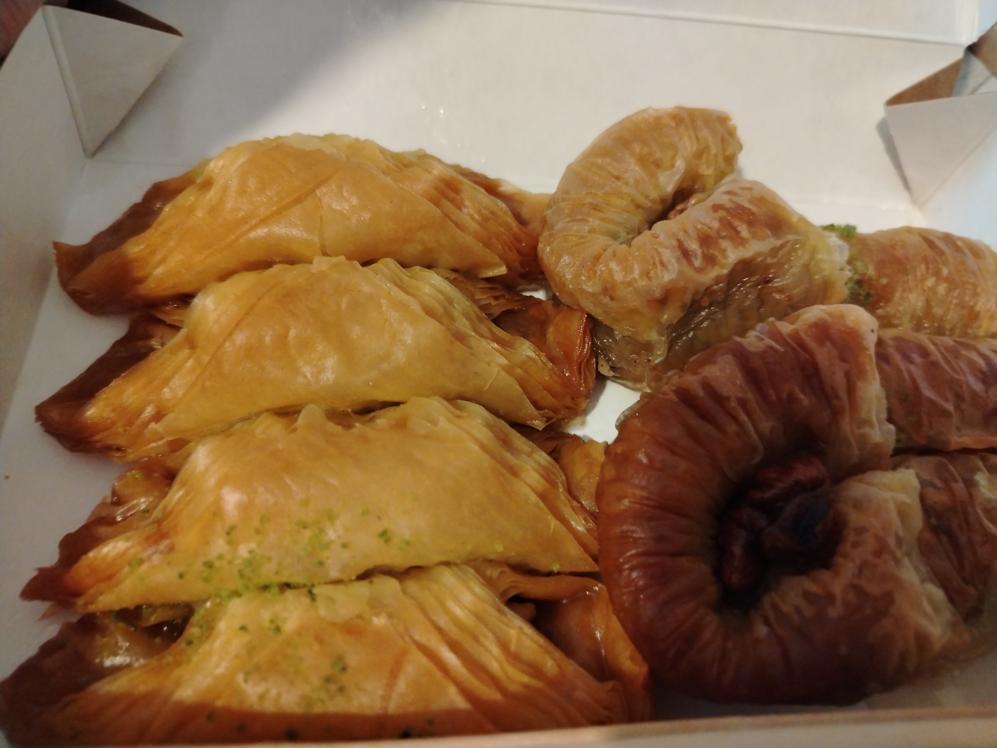 A close-up shows an assortment of baklava pastries inside a white cardboard box. There are several triangular-shaped pieces, some sprinkled with green pistachio crumbs. Next to these are rolled, spiral pastries. A round pastry with a dark center is also present. The pastries have a glossy, honey-colored appearance. The setting appears to be indoors, with the box sitting on a flat surface. There is no indication of the city of Noviy, Russia in the image.