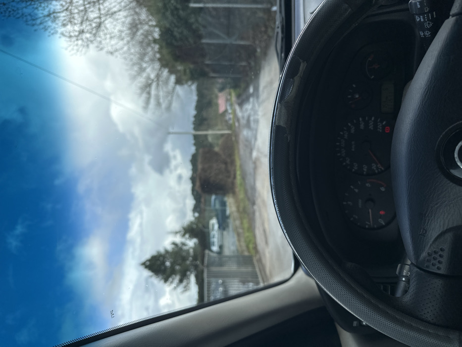 The image depicts a first-person perspective from inside a stationary vehicle in Vila Real, Portugal, looking out through the windshield. The vehicle's steering wheel, partially covered by a distressed, peeling black and grey cover, occupies the lower portion of the frame. The dashboard instrument cluster is visible, showing a tachometer on the left and a speedometer (up to 220 km/h) on the right, with several illuminated warning lights.
Beyond the windshield, the scene shows an unpaved, wet, and muddy track extending into the distance under an overcast sky with patches of blue and bright sunlight. To the left of the track, a metal gate leads to an area containing several parked vehicles, including light-colored vans, behind which a tall evergreen tree is prominent. To the right of the track, a trimmed, dark green hedge or large bush is present, followed by a metal fence and bare deciduous trees. A utility pole stands further down the track on the right. In the far background, a building with a reddish roof is faintly visible amidst more distant foliage. No people are visible.