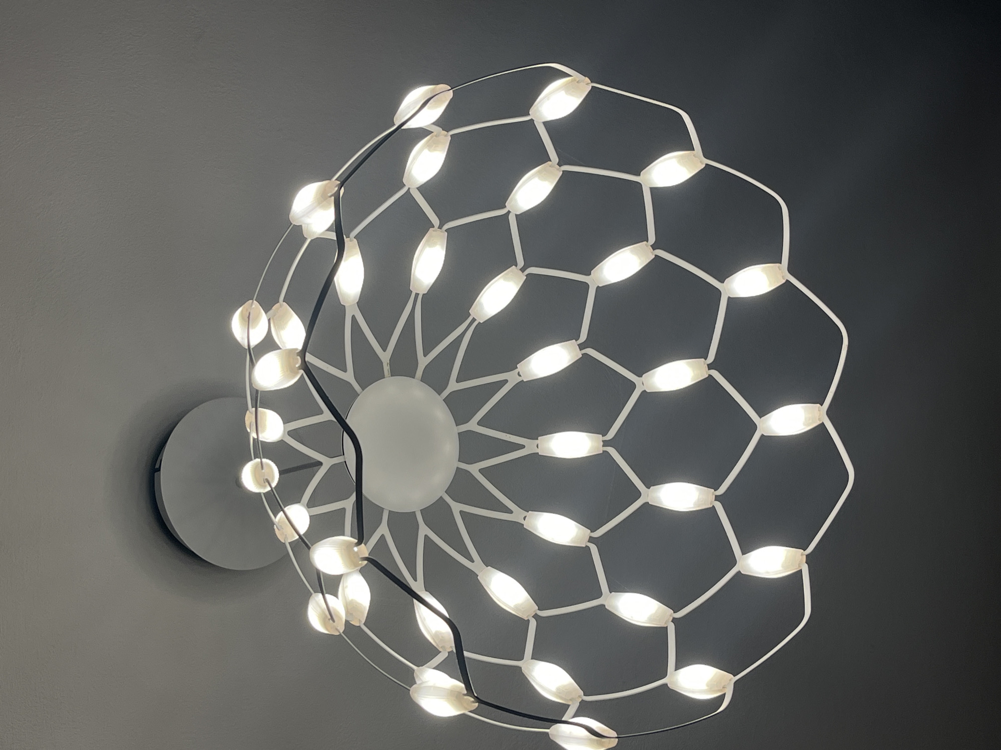 A modern white chandelier hangs against a gray wall. The chandelier consists of a circular arrangement of connected white hexagonal frames, with an elongated white bulb at each joint. A black wire snakes through the chandelier's structure. The mounting fixture is a circular disk, also white. The scene appears to be indoors.