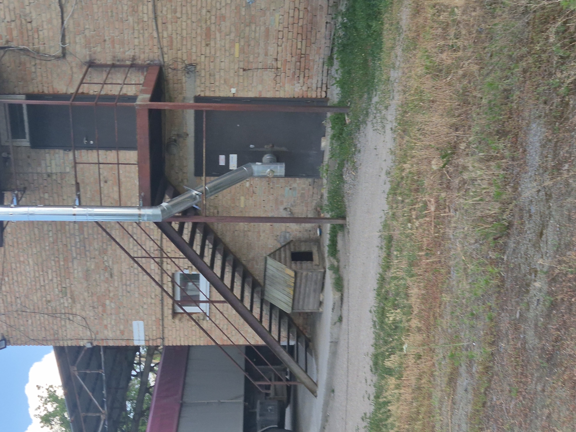 The image shows the exterior of a brick building in Sozonivka, Ukraine. A metal external staircase with a rusted railing ascends to a landing with a dark door and a balcony. A large metal pipe runs vertically along the side of the building, connected to a smaller pipe that angles downwards. Below the staircase, a small wooden doghouse is visible on a concrete surface. To the left of the building, the side of a large truck with a red tarp is visible. The ground in the foreground is a mix of gravel and dry grass. The sky is visible in the upper left corner.