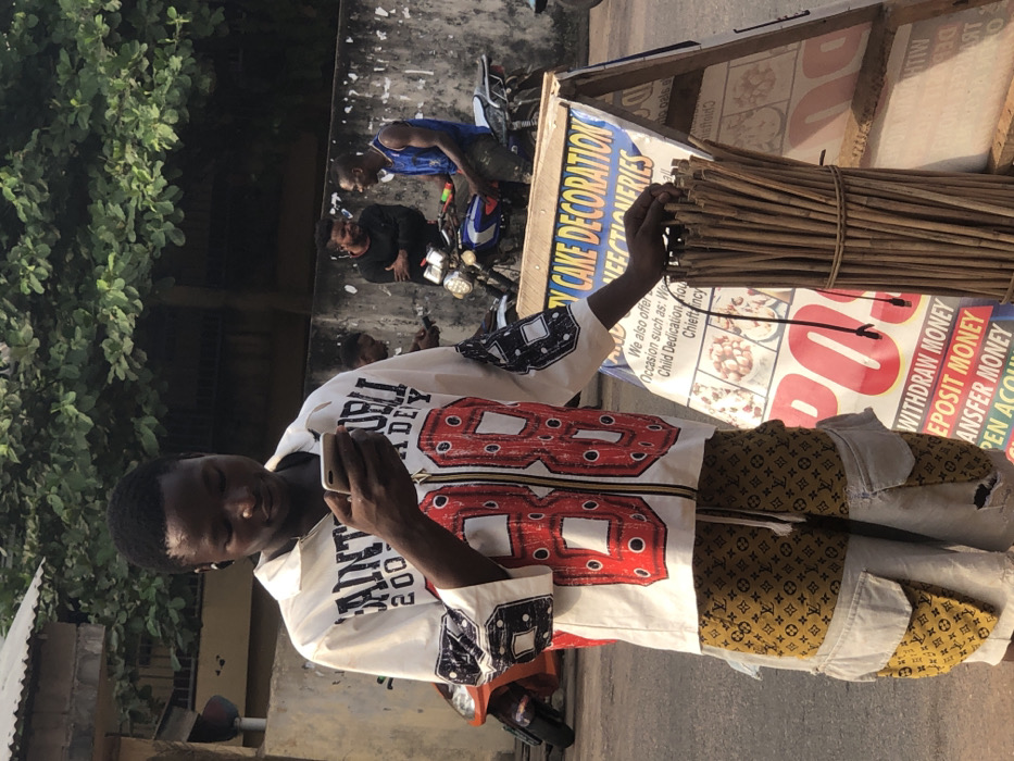A man is looking at a cell phone and standing in front of a sign that reads "TY CAKE DECORATION." The sign also advertises withdrawal, deposit, and transfer money. The man is wearing a white shirt with a large red "88" on the front and gold and brown patterned pants. In the background, there are other people and motorbikes. Green foliage is visible on the left. The scene is outdoors, and the lighting suggests it is daytime.