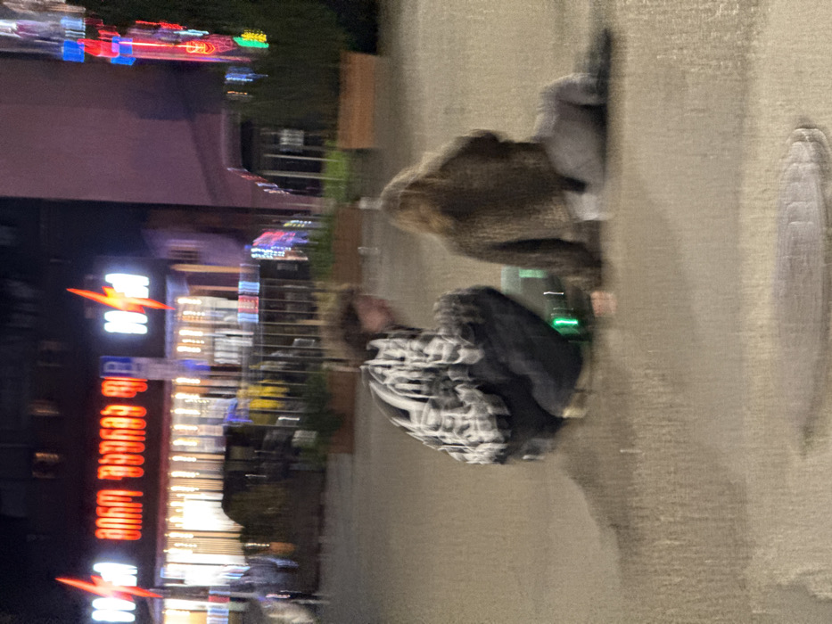 The image depicts a blurred, nighttime urban scene on a wet, paved surface, likely after rain. In the foreground, two individuals are positioned on the ground, interacting with a small, illuminated green object between them. The person on the left is squatting, facing right, wearing a patterned, light-colored long-sleeved shirt and dark trousers. The person on the right is seated, facing left, with light-colored hair, wearing a dark, textured jacket and light-colored trousers.
The background features a building with prominent, illuminated signage. A large red sign displays Georgian script "შიმ ქიმ" (Shum Kim), flanked by two distinctive red lightning bolt logos, characteristic of a fast-food restaurant. A "P" parking sign is also visible. This establishment identifies the location as Tbilisi, Georgia. To the far right, a vertical structure with multiple colored lights (red, blue, green) adds to the urban illumination. The overall scene is dimly lit, with reflections on the wet ground surface.
