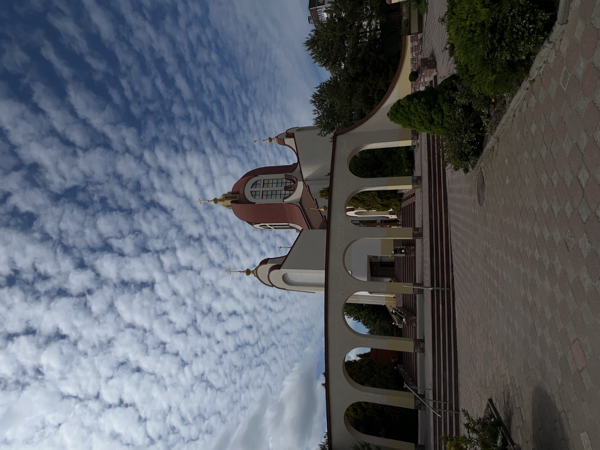 The image shows the Cathedral of the Ternopil Diocese of the Ukrainian Orthodox Church in Ternopil, Ukraine. It is a white building with a red roof and domes. The building has a series of arches along one side. The sky is blue with a pattern of small white clouds. A brick path is in the foreground.
