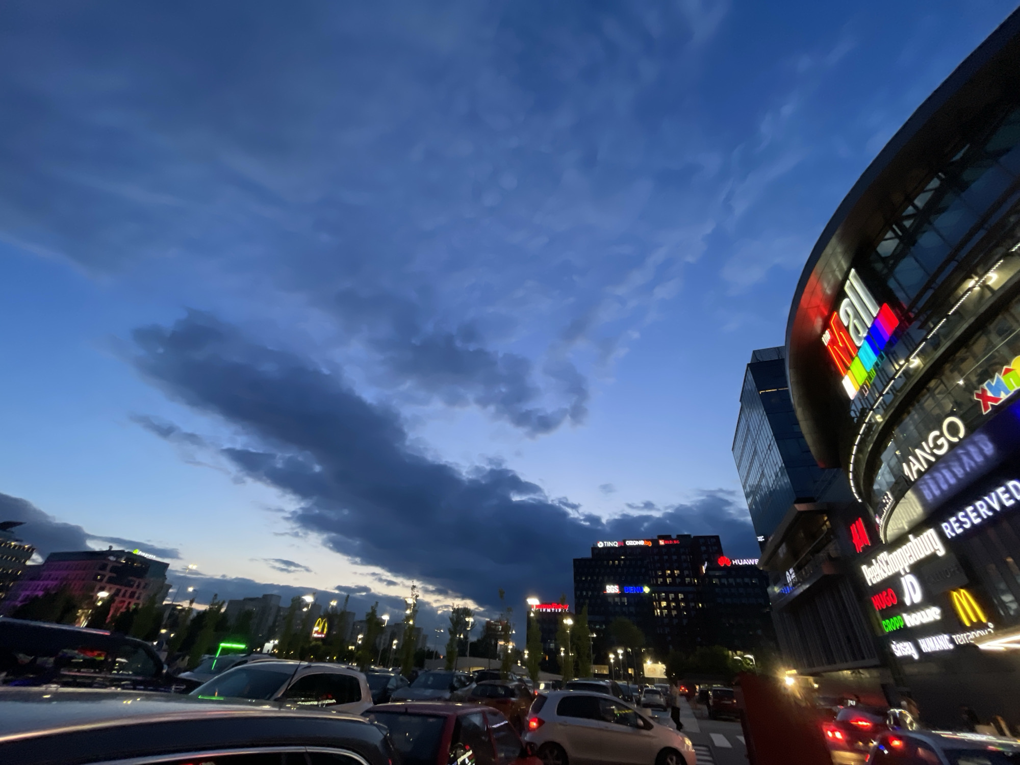 The photograph captures a cityscape scene at dusk. Dominating the right side of the frame is a large, modern shopping mall identified by the illuminated letters "MALL" in a vibrant array of colors, with notable brand signage including MANGO, HUGO, and McDonald's. Adjacent to the mall, a tall, slender glass building can be seen. In the foreground, a parking area is filled with numerous cars of various makes and models, indicating a busy location. Across from the mall and behind the parking area, a building with illuminated brand logos on its roof is visible. The sky is filled with dark, dramatic clouds tinged with hints of sunset, and streetlights illuminate the area, further contributing to the twilight ambiance.