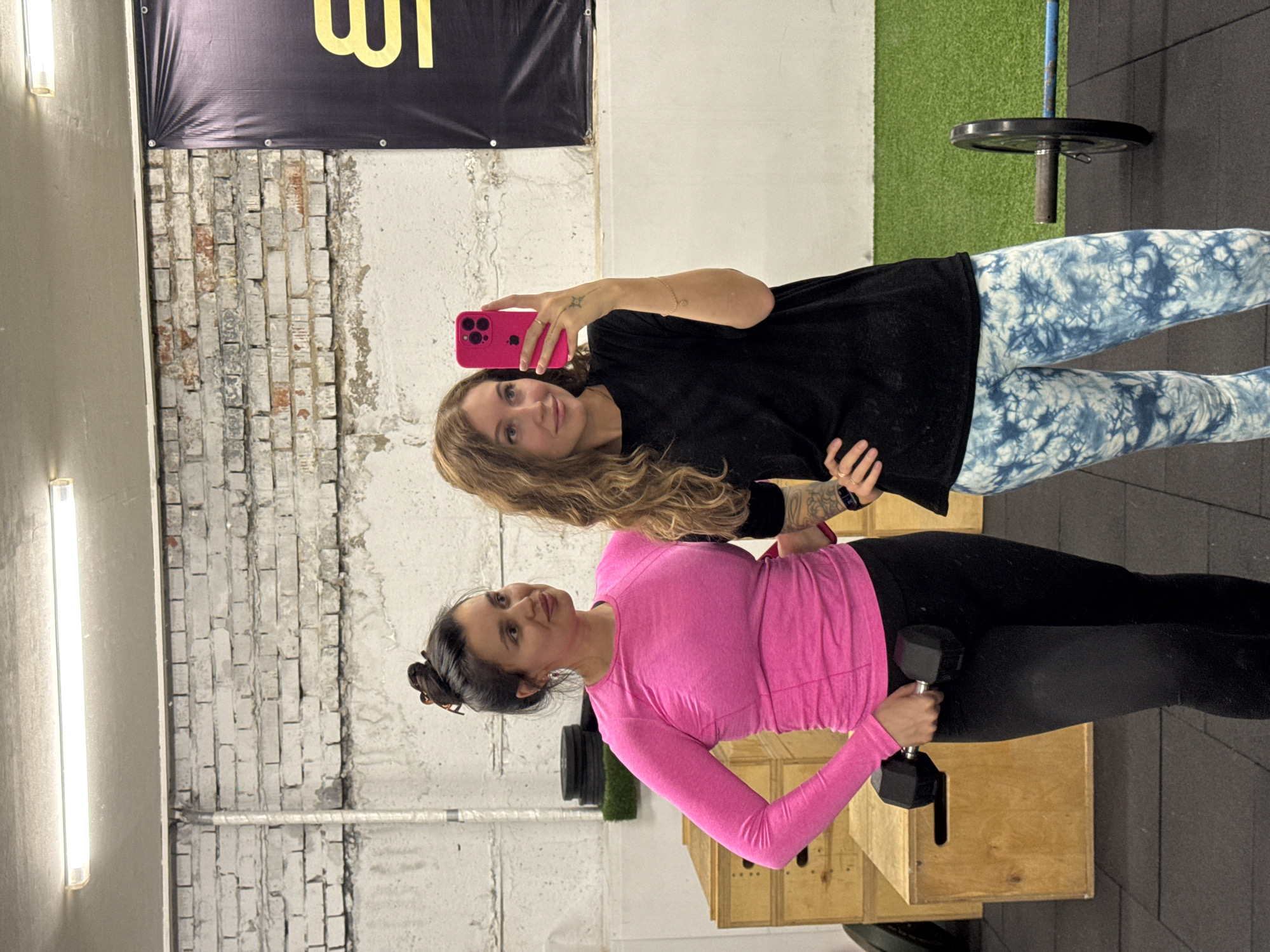 A portrait-oriented photo captures two women inside what appears to be a gym or fitness studio in Chaikovsky, Russia. The setting features raw, industrial-style walls with exposed, light-colored brickwork contrasting with painted concrete, illuminated by a long fluorescent light fixture. The floor is covered with black rubber gym mats and a strip of green artificial turf.
On the right, a woman with long, wavy blonde hair is taking a mirror selfie using a bright pink smartphone. She wears a black long-sleeved top and distinctive blue and white tie-dye leggings, and has a small tattoo visible on her right wrist. She is looking directly at the camera with a faint smile.
On the left, another woman with dark hair pulled back into a bun is holding a black, hexagonal dumbbell in her right hand. She is dressed in a long-sleeved, bright pink athletic top and black leggings. She stands next to a wooden plyometric box, with a barbell and weight plate partially visible behind her. Her expression is neutral and focused.
In the upper background, a black banner with partial yellow text, possibly Cyrillic characters like "ЦІ" or "УГІ", is visible. The scene suggests a casual fitness session, possibly a workout or a break from one, captured in a selfie moment.