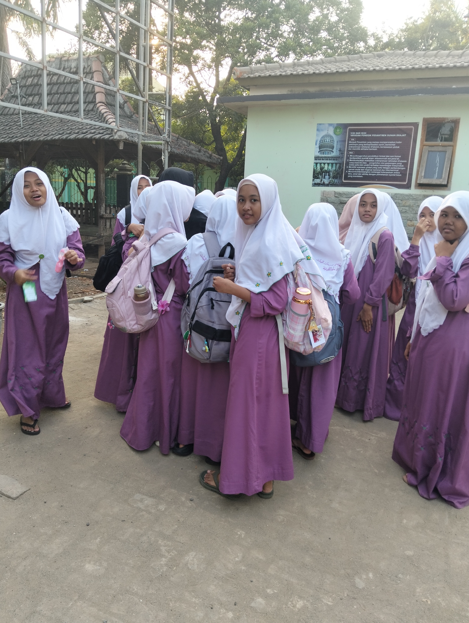 In the city of Paciran, Indonesia, a group of young Muslim girls, all wearing matching purple robes and white hijabs, are gathered outdoors. Some of the girls are carrying backpacks, suggesting they are students. The background includes a light green building with a sign displaying text and an image of a domed structure. There's also a glimpse of a covered outdoor structure with a tiled roof and metal framework, as well as trees and foliage. The ground is a dirt or gravel path. The overall scene appears to be a school or religious institution campus.