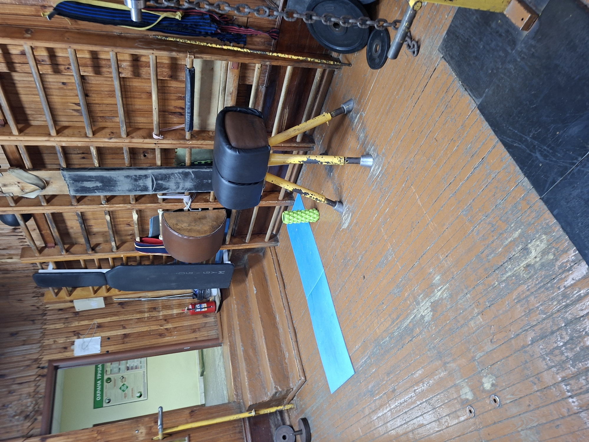 The image depicts a gym with wooden walls and floor. On the floor, a blue yoga mat is unrolled, next to a green foam roller. In the center of the image is a pommel horse with a black leather top and yellow legs. Behind the pommel horse are wooden shelves with various gym equipment, including a long black mat, a brown barrel-shaped object, and a chain. To the left, a fire extinguisher is visible next to a set of wooden stairs leading to a doorway. The doorway opens to a room with a poster on the wall that reads "ОХРАНА ТРУДА" (Labor Protection). The overall scene suggests a functional but somewhat worn gym environment.