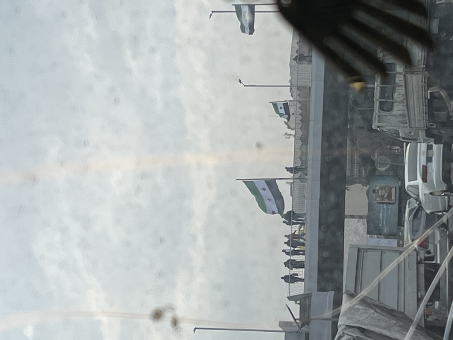 The image displays an outdoor scene in Al-Muhajerin Municipality, Syria, captured from within a vehicle looking towards a concrete overpass or bridge under an overcast sky. Approximately 8-12 individuals are present on the overpass; most are standing in small groups near the center, while at least two figures are walking towards the right. Three flags, identifiable as the Syrian opposition/revolution flag (green, white, and black horizontal stripes with three red stars in the white band), are prominently displayed on poles along the overpass. Streetlights are visible on and adjacent to the overpass. Below the overpass, a road with vehicular traffic includes a white sedan and parts of two trucks. A large billboard featuring text and an image is visible beneath the overpass. The photograph contains numerous circular dust or dirt artifacts and a vertical light streak, suggesting it was taken through a soiled window or lens. A hand is partially visible in the lower right foreground, obscuring a portion of the scene.