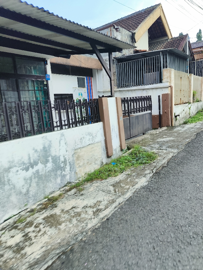 The image shows a street in Bandung, Indonesia, located at coordinates -6.895 latitude and 107.653 longitude. The street is paved with asphalt and runs along a white concrete wall with a brown concrete pillar. The wall has a metal fence on top of it and a small window. A brown wooden gate is on the right side of the wall. There is a small patch of grass in front of the gate. On the other side of the street is a building with a red tile roof. The building has a metal fence on the front and a wooden door. The image was taken from a perspective looking down the street. There are no people in the image.