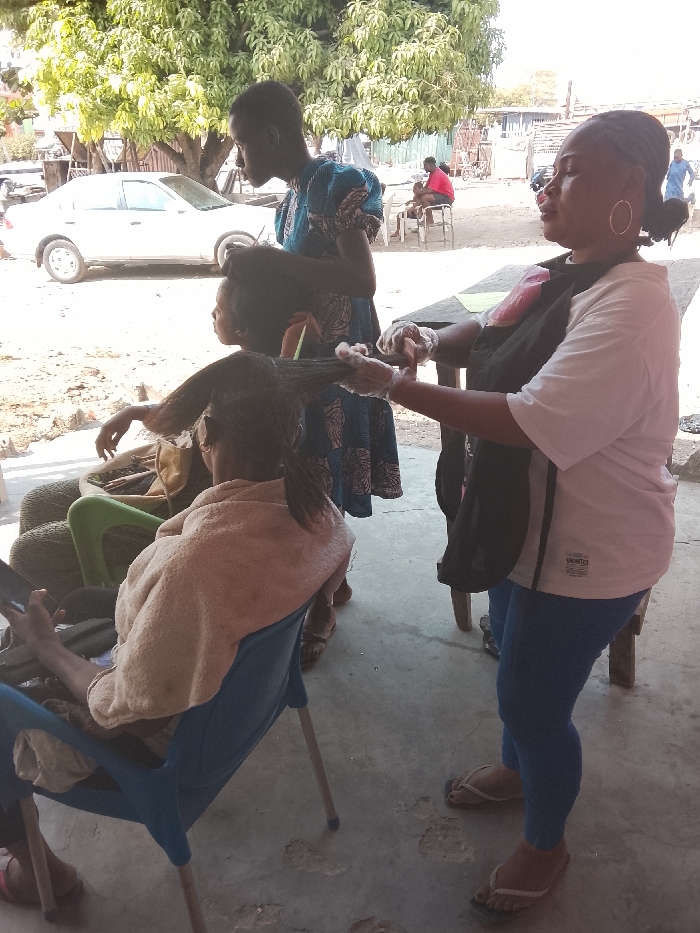 The image depicts an outdoor or semi-outdoor scene during bright daylight, likely a hair salon or styling area. In the foreground, three individuals are engaged in a hair styling activity.
On the far left, seated in a blue plastic chair with their back to the camera, is a person whose hair is being treated. They are draped with a light-colored (possibly beige or light brown) towel or shawl over their shoulders. Their long, dark hair is pulled back and sectioned, heavily coated with a thick, greenish-brown product, suggesting a chemical treatment like a relaxer, dye, or deep conditioner. This person holds a mobile phone in their left hand, appearing relaxed while waiting. Behind them, several stacked tires are visible, possibly serving as additional seating or part of the outdoor setup.
Standing to the right of the seated individual and facing left is a woman actively working on the hair. She wears a white short-sleeved t-shirt with small text visible on the front (partially readable as "UNLIMITED"), blue leggings, and light-colored flip-flops. She has a dark apron or bib covering her front and is wearing clear plastic gloves on both hands as she applies the product to the client's hair with focused attention. Her hair is pulled back, and she wears large hoop earrings.
Between the seated client and the standing stylist, a young person, possibly a child or teenager, stands observing or assisting. They are wearing a blue patterned dress and have short, dark hair, looking down at the hair being worked on.
In the mid-ground, a white car is parked on the left side, partially obscured by a large, leafy green tree. Further in the background on the right, another person in a red top is visible seated at a table, along with other blurry figures, suggesting a bustling outdoor environment. The ground is a light-colored concrete or paved surface, showing some wear and cracks in places. The bright sunlight indicates a clear day, casting soft shadows.