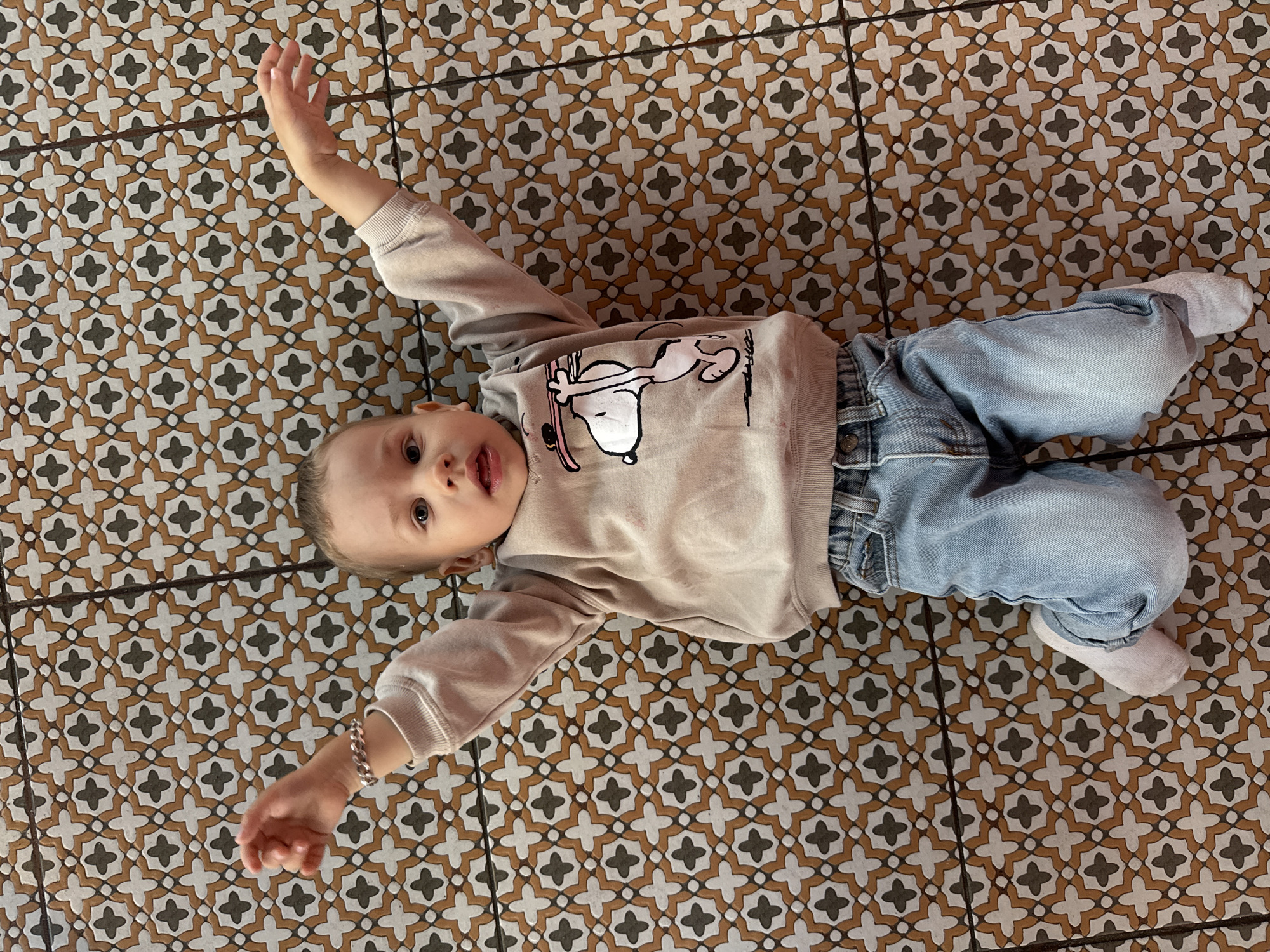 A baby wearing a beige sweater with Snoopy on it and jeans is lying on a patterned tile floor. The baby is lying on their back with their arms outstretched to their sides. The background is a tile floor with a pattern of black and white crosses on a brown background.