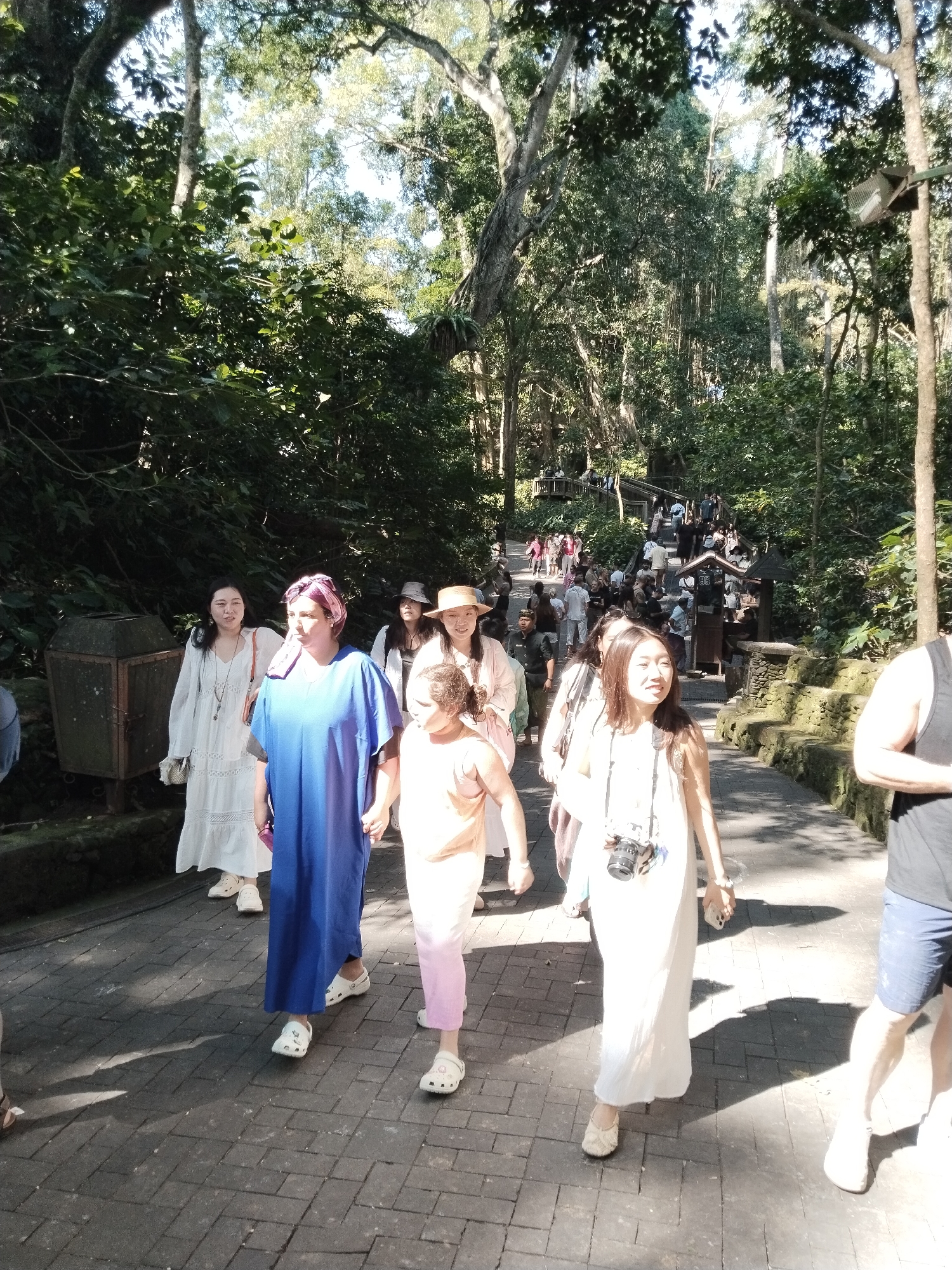 In Ubud District, Indonesia, a group of people are walking along a paved path surrounded by lush greenery. The path is made of dark, rectangular stones, with some moss growing in the crevices. Sunlight filters through the dense canopy of trees, casting dappled shadows on the ground.
In the foreground, several individuals are prominent. A woman in a long blue dress and a purple headscarf is walking with her hands clasped. To her left, a woman in a white flowing dress with intricate embroidery on the skirt smiles at the camera, holding a straw bag. To the right of the woman in blue, a young girl in an orange top and pink pants walks hand-in-hand with another woman. This woman is wearing a white dress and has a camera around her neck. Further ahead, other people are visible, some walking in the same direction, others interacting. In the background, the path curves upwards, leading to a more densely populated area with many more people, suggesting a popular tourist destination. A trash bin made of metal is visible on the left side of the path. The overall impression is one of a lively scene in a tropical environment.