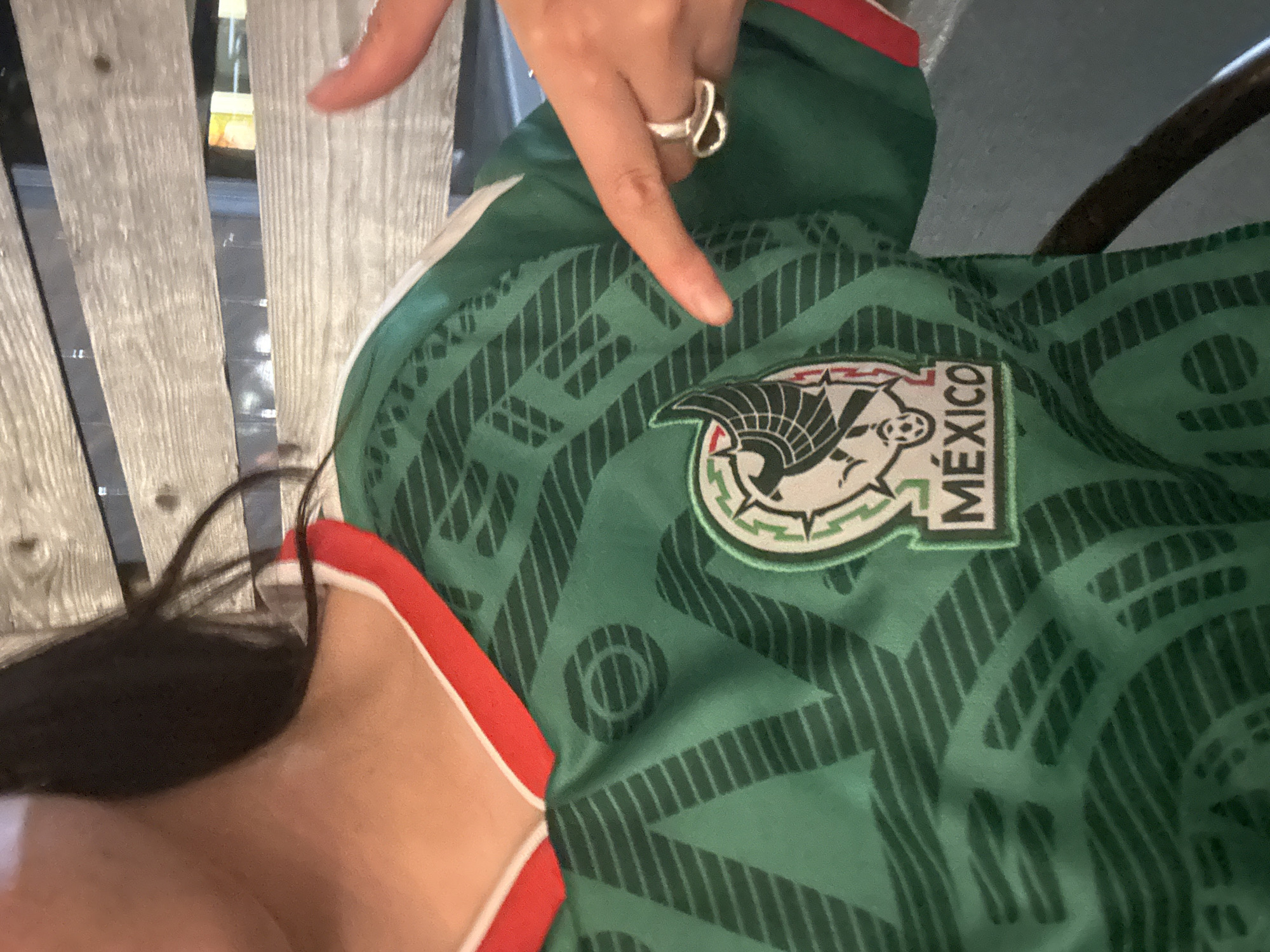 A close-up shot shows a person wearing a green and white Mexico soccer jersey. The jersey has a red trim around the collar and sleeves. The person's right hand is visible, pointing towards the national emblem on the jersey. The emblem is circular with a stylized eagle and serpent, and the word "MÉXICO" is printed below it. The background consists of wooden slats, suggesting an outdoor or rustic indoor setting. The image is taken from a mid-chest view, focusing on the upper body and the jersey.