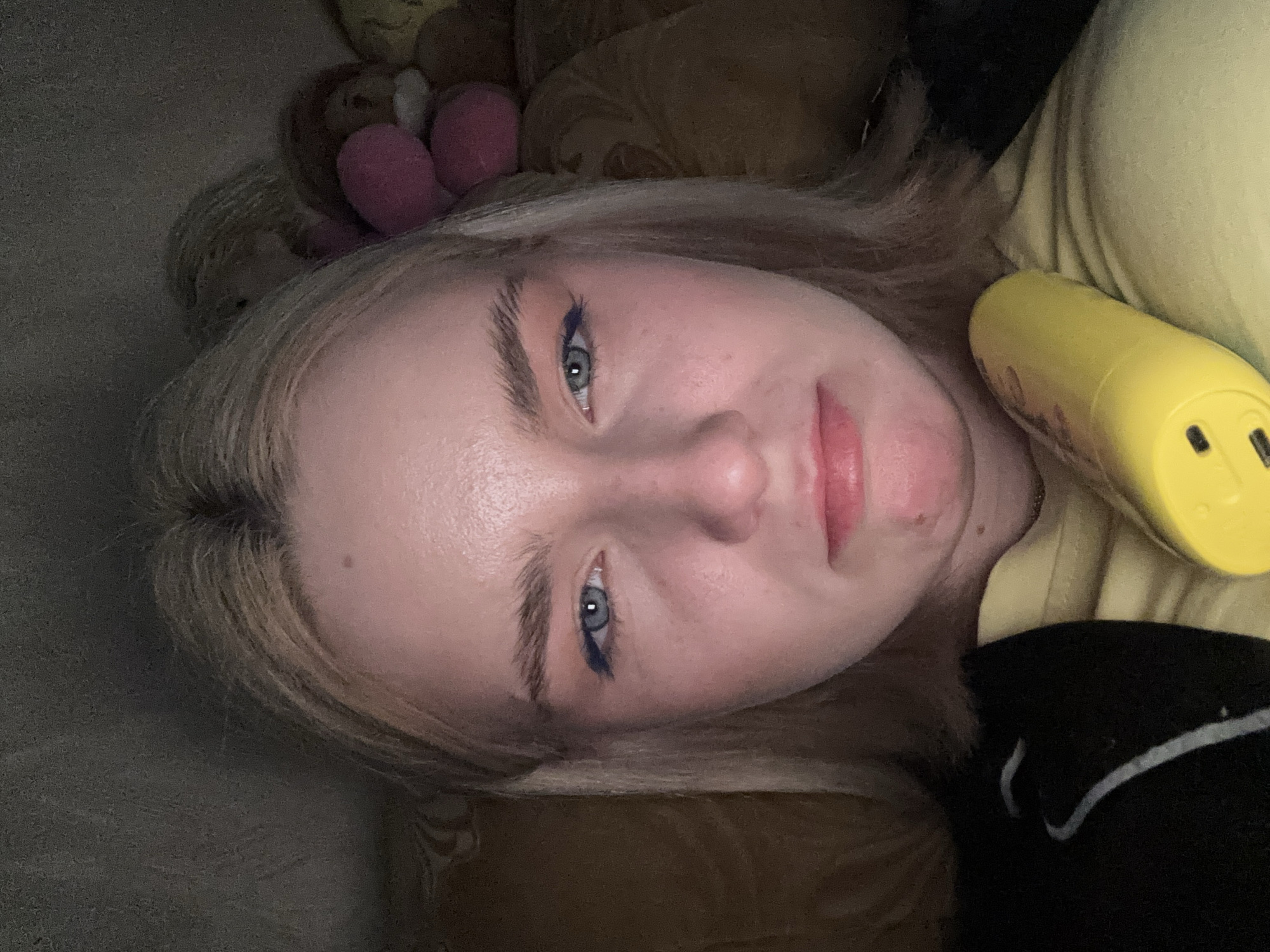 The image is a close-up, vertically oriented, selfie-style photograph of a young person with blonde hair. The person is lying down, and their face is tilted towards the camera. They have light blue eyes and are wearing dark blue eyeliner. Their lips are slightly parted, and their skin appears to be pale with some subtle redness on the cheeks.
In the background, there are what appear to be stuffed animals, including one with pink pom-pom-like features and another that is brown and fuzzy. To the right of the person's face, a bright yellow cylindrical object is visible, which might be a bottle or a portable charger, with what looks like USB ports. The person is wearing a light yellow garment on their upper body. The lighting is dim, suggesting it might be nighttime or a poorly lit room. The overall mood appears relaxed or casual. There is no visible text in the image.