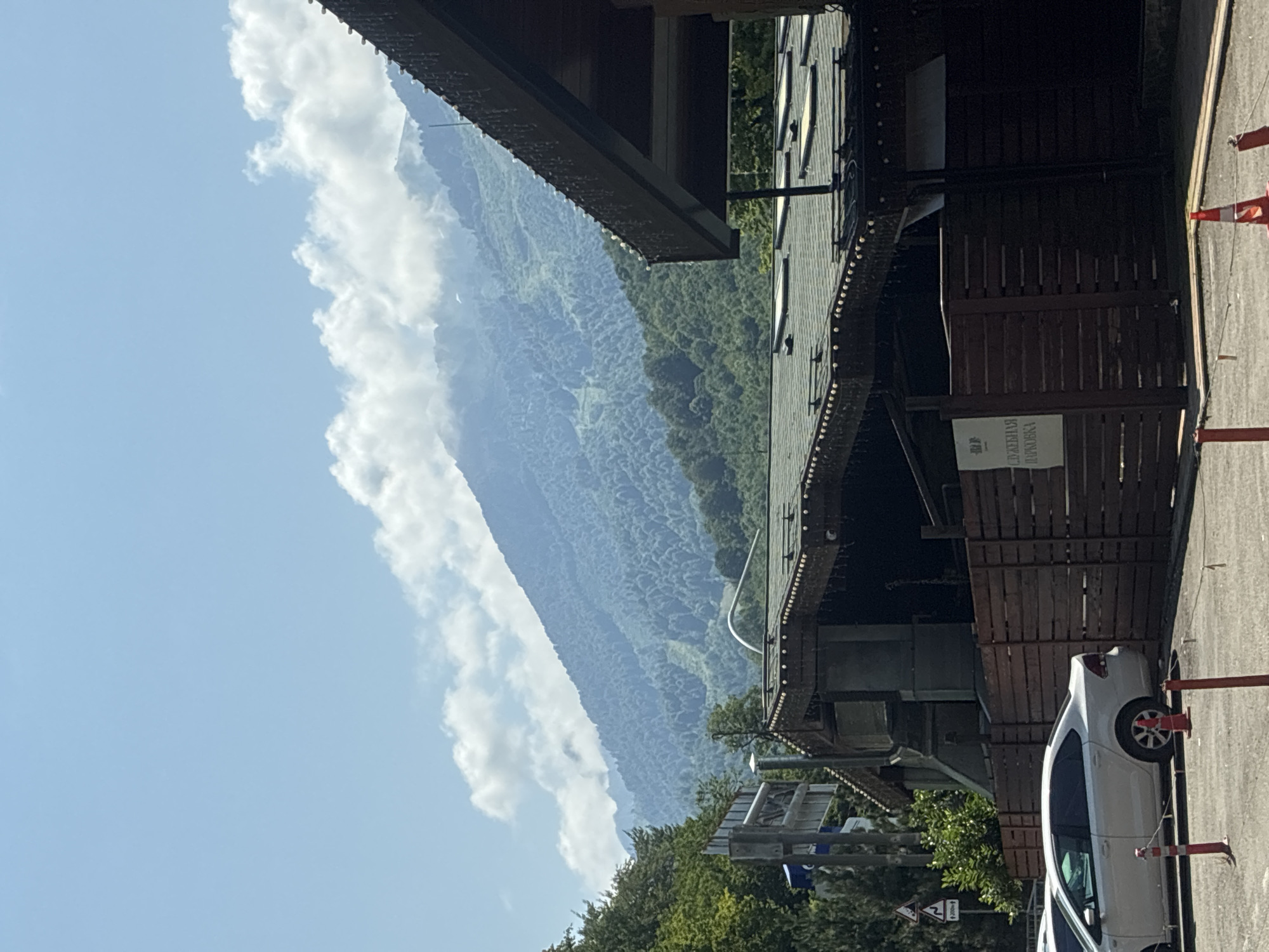 In the city of Estosadok, Russia, a view captures a vast mountain range covered in dense green forest and partially obscured by clouds under a clear blue sky. In the foreground, a wooden building with a dark roof, adorned with decorative lights, stands prominently. A wooden fence borders the area. A white sedan is parked in front of the fence, with traffic cones and bollards visible in the foreground. A sign with Russian text, indicating "SERVICE PARKING," is affixed to the fence. The overall scene depicts a serene, mountainous landscape juxtaposed with man-made structures.