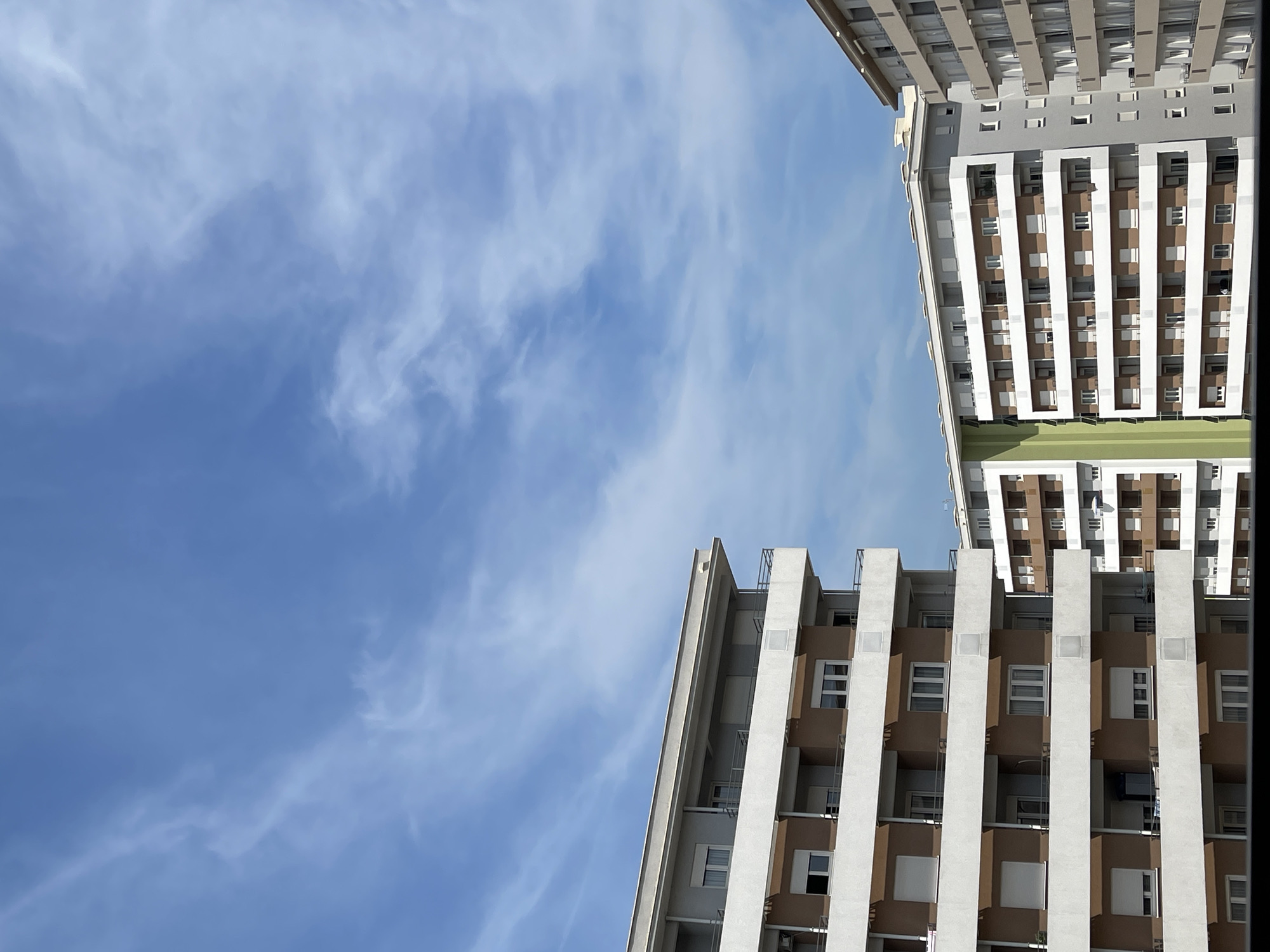The image shows a view of apartment buildings against a blue sky with wispy clouds. The buildings are modern, with multiple stories and balconies. The color palette is dominated by neutral tones of white, beige, and brown, with a vertical strip of green visible on one of the buildings. The scene appears to be a residential area within a city, potentially Podgorica, Montenegro, characterized by urban architecture and a clear sky. There are no people or specific actions depicted, focusing solely on the architectural structures and the atmospheric conditions.