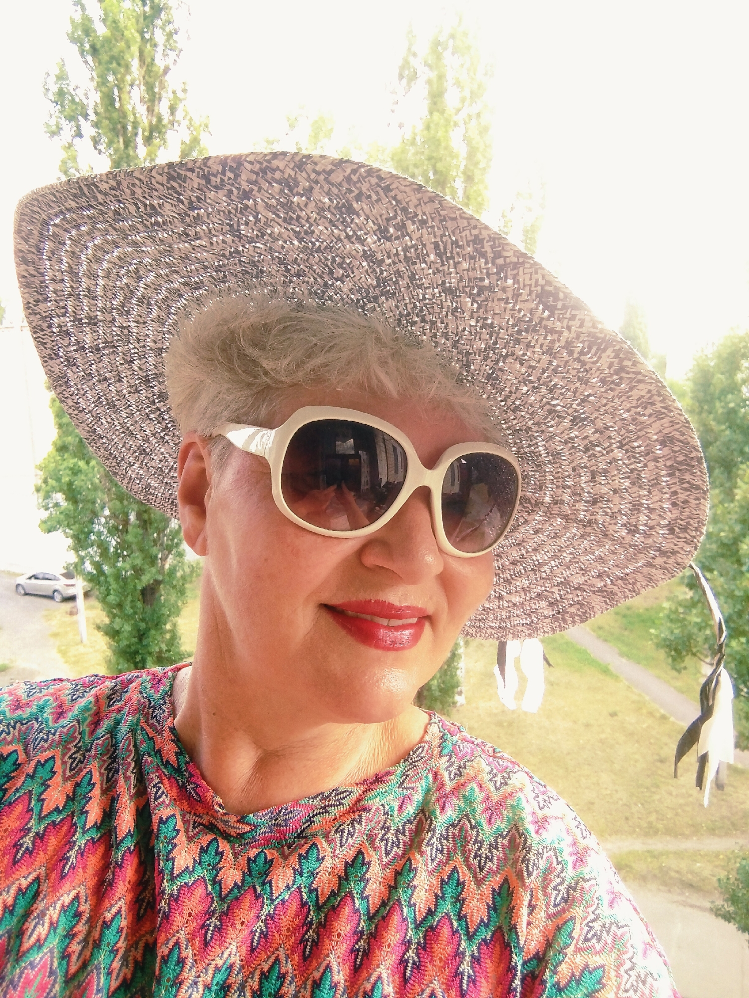 The image is a close-up, selfie-style shot of a woman wearing a wide-brimmed straw hat and large, white-rimmed sunglasses. She has short, light-colored hair and is smiling, showing her teeth, with red lipstick. Her shirt features a colorful, intricate zigzag pattern in shades of pink, orange, green, and blue. The background is blurred but appears to be an outdoor setting with trees and a faint view of a street with a silver car, suggesting an urban or suburban environment. The overall lighting indicates it is daytime, possibly sunny, with some overexposure in the upper portion of the image. The image was taken from a slightly elevated position, looking down at the subject. The presence of trees and buildings indicates a location within the city of Horishni Plavni, Ukraine.