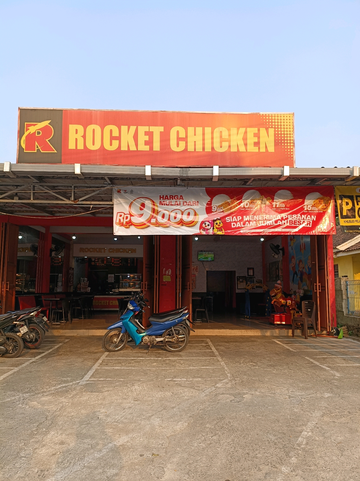 The image shows the exterior of a "Rocket Chicken" restaurant located in Ngemplak, Indonesia. The restaurant has a prominent red and yellow sign that reads "ROCKET CHICKEN" with a stylized rocket logo. Below the sign, a banner displays "HARGA MULAI DARI RP 9.000" (Prices starting from RP 9,000) and "SIAP MENERIMA PESANAN DALAM JUMLAH BESAR" (Ready to accept large orders). The entrance to the restaurant is visible, with glass doors and red-painted pillars. Inside, tables and chairs are set up, and a counter area is visible. In front of the restaurant, several motorcycles are parked in marked bays on a concrete lot. A bright blue motorcycle is in the foreground, parked on the right side of the frame. The environment appears to be an outdoor, daytime setting with a clear sky.