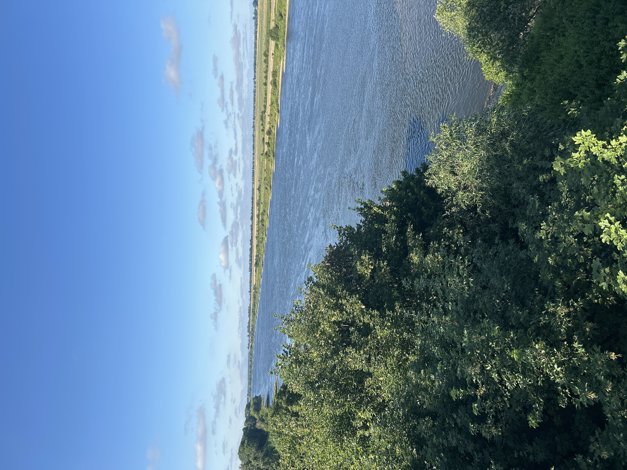 A wide, deep river flows through a rural landscape under a bright blue sky dotted with white clouds. Lush green trees and foliage line the foreground on the left bank of the river, with a gently sloping embankment visible. The opposite bank features cultivated fields stretching towards the horizon, with a thin line of trees marking the distant boundary. The water's surface is rippled, reflecting the sunlight. In the far distance, faint outlines of structures or wind turbines are visible against the horizon. The scene captures a serene and natural setting in Bałdowo, Poland, likely during daylight hours in the summer or late spring.