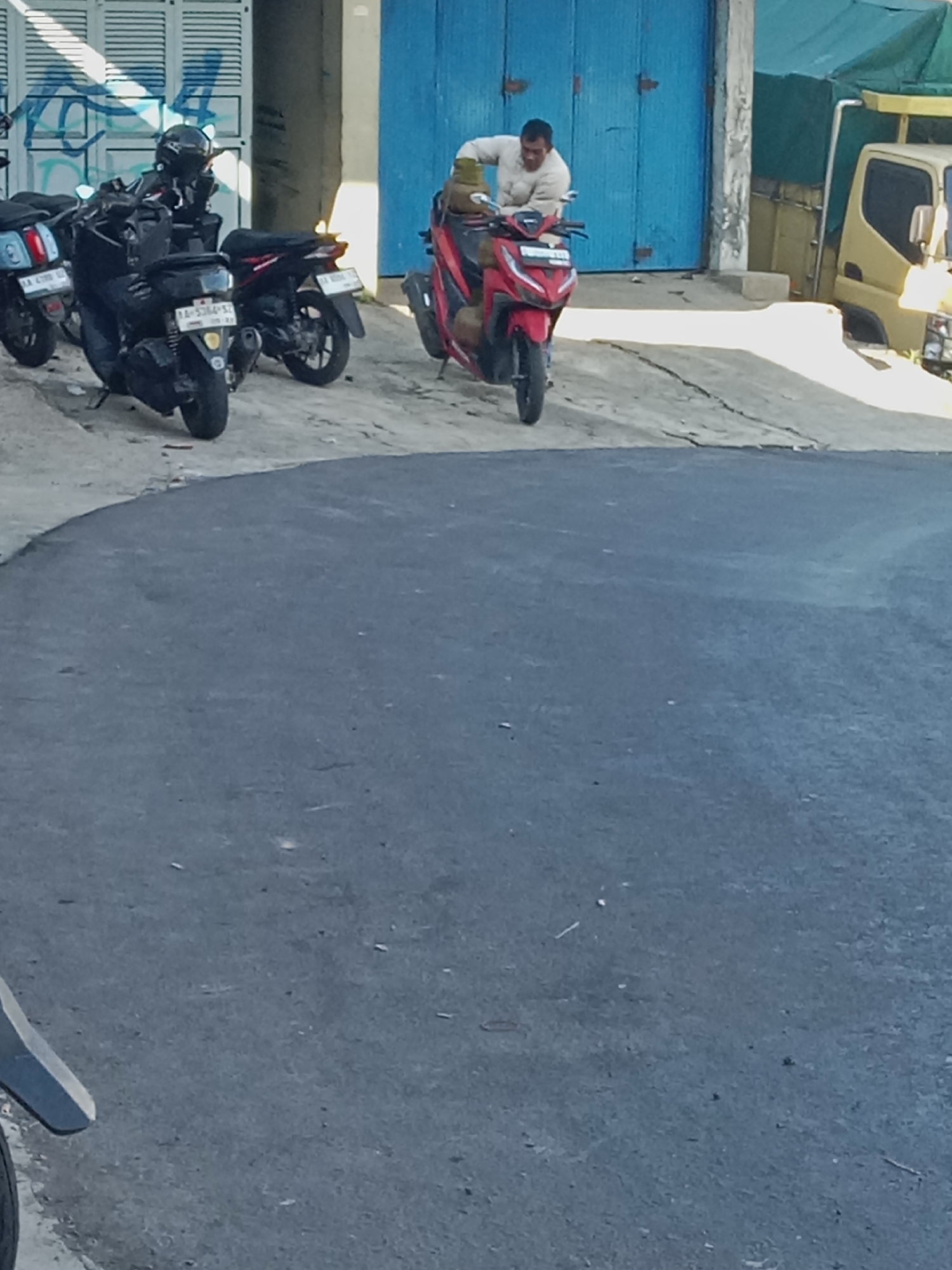 In Kejajar, Indonesia, a man in a light-colored shirt is riding a red motorcycle past a building with a large blue door. The motorcycle is in motion, and the man is leaning forward. Several other motorcycles are parked nearby. A yellow truck is visible in the background. The scene appears to be on a street or walkway with cracked pavement.