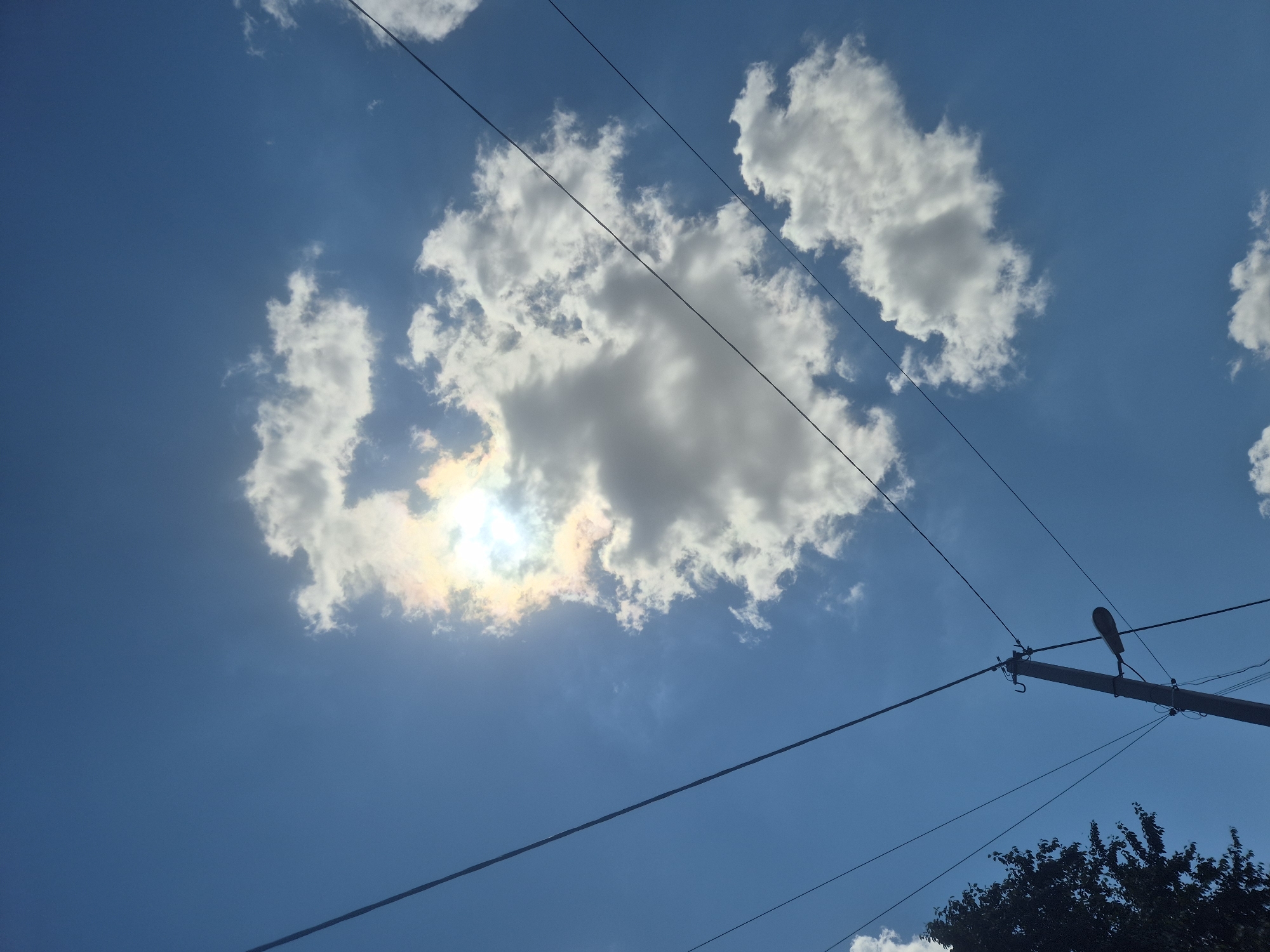 The image captures a bright, sunny day in Sozonivka, Ukraine, as evidenced by the clear blue sky and scattered white clouds. A prominent cloud formation is visible, partially obscuring the sun, which creates a rainbow-like effect. Power lines stretch across the frame, indicating an urban or suburban setting. In the lower left portion of the image, a portion of a dark green tree is visible, adding a touch of natural scenery. A utility pole with a street lamp and several wires is also present, suggesting infrastructure supporting the community. No people are visible in the image. The overall impression is one of a typical day, with a focus on the sky and the surrounding environment.